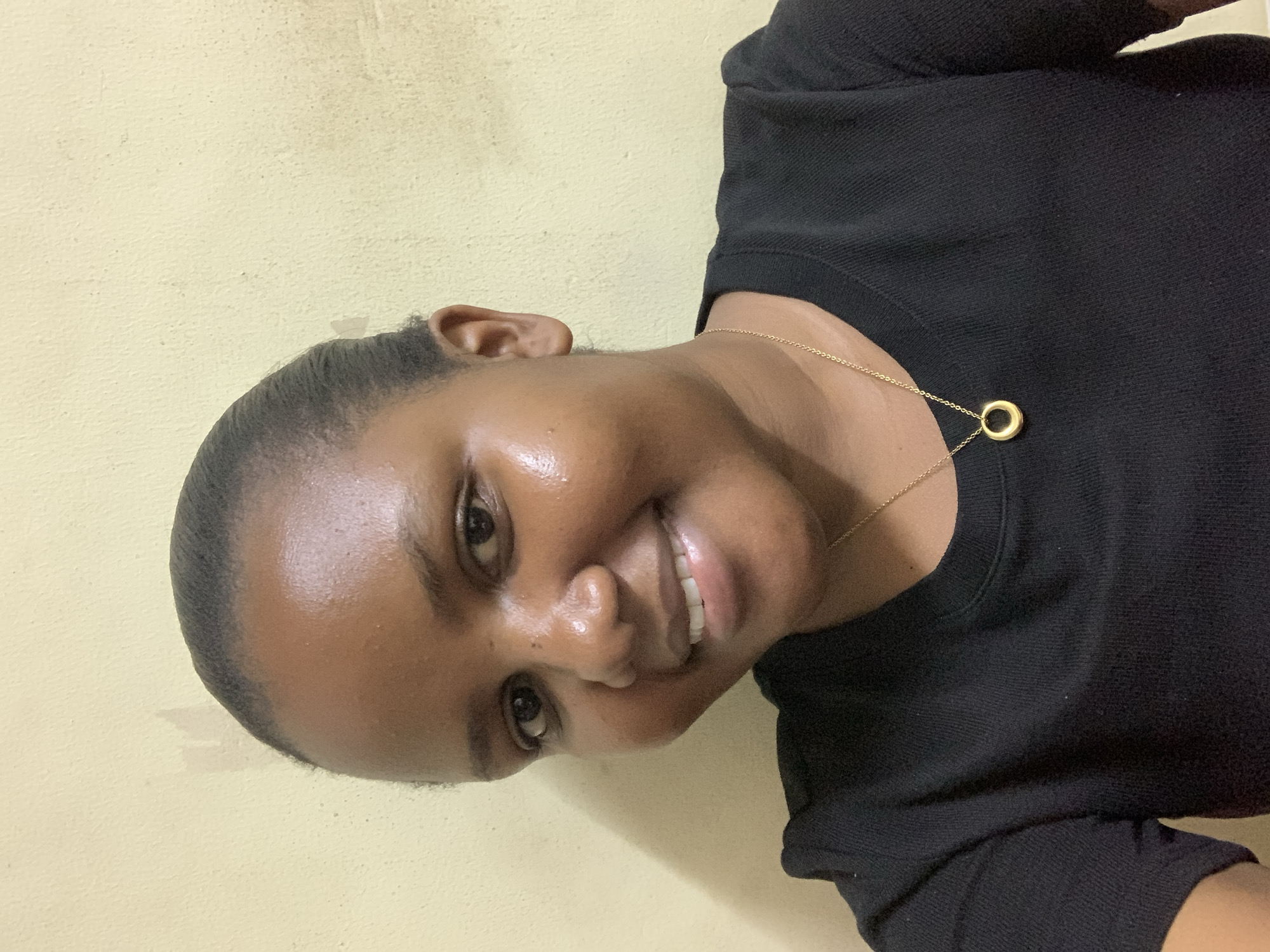 A medium shot captures a woman with short, dark hair pulled back. She has medium brown skin and is smiling, showing her teeth. She wears a black top and a gold necklace with a circular pendant. The background is a light yellow, slightly textured wall. The location is not discernible from the visual information provided in the image. There is no indication it is in Dar es-Salaam, Tanzania.