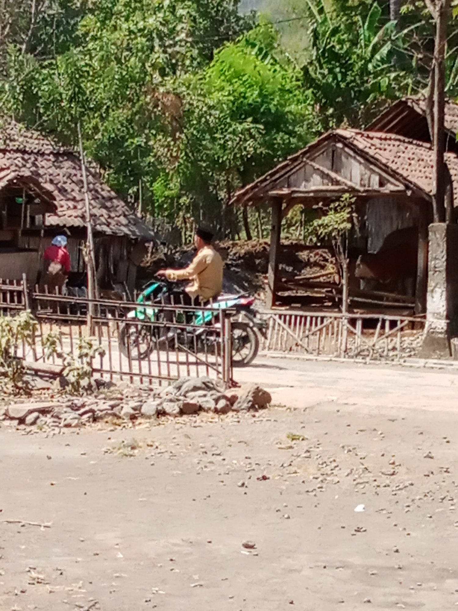 In the city of Selowogo, Indonesia, a person wearing a light brown long-sleeved shirt and a black cap is seated on a teal and white motorcycle. The motorcycle has a silver exhaust pipe and black tires. The person is on a dusty, unpaved road, with their left hand on the motorcycle's handlebar. To the left of the motorcycle, there is a dark metal fence. Behind the fence, another person in a red shirt and a blue head covering is standing near a rustic, thatched-roof structure. The structure is made of wood and has a tiled roof. Further in the background, lush green trees and vegetation dominate the landscape, with the hint of a hill or mountain. The scene is illuminated by bright sunlight.