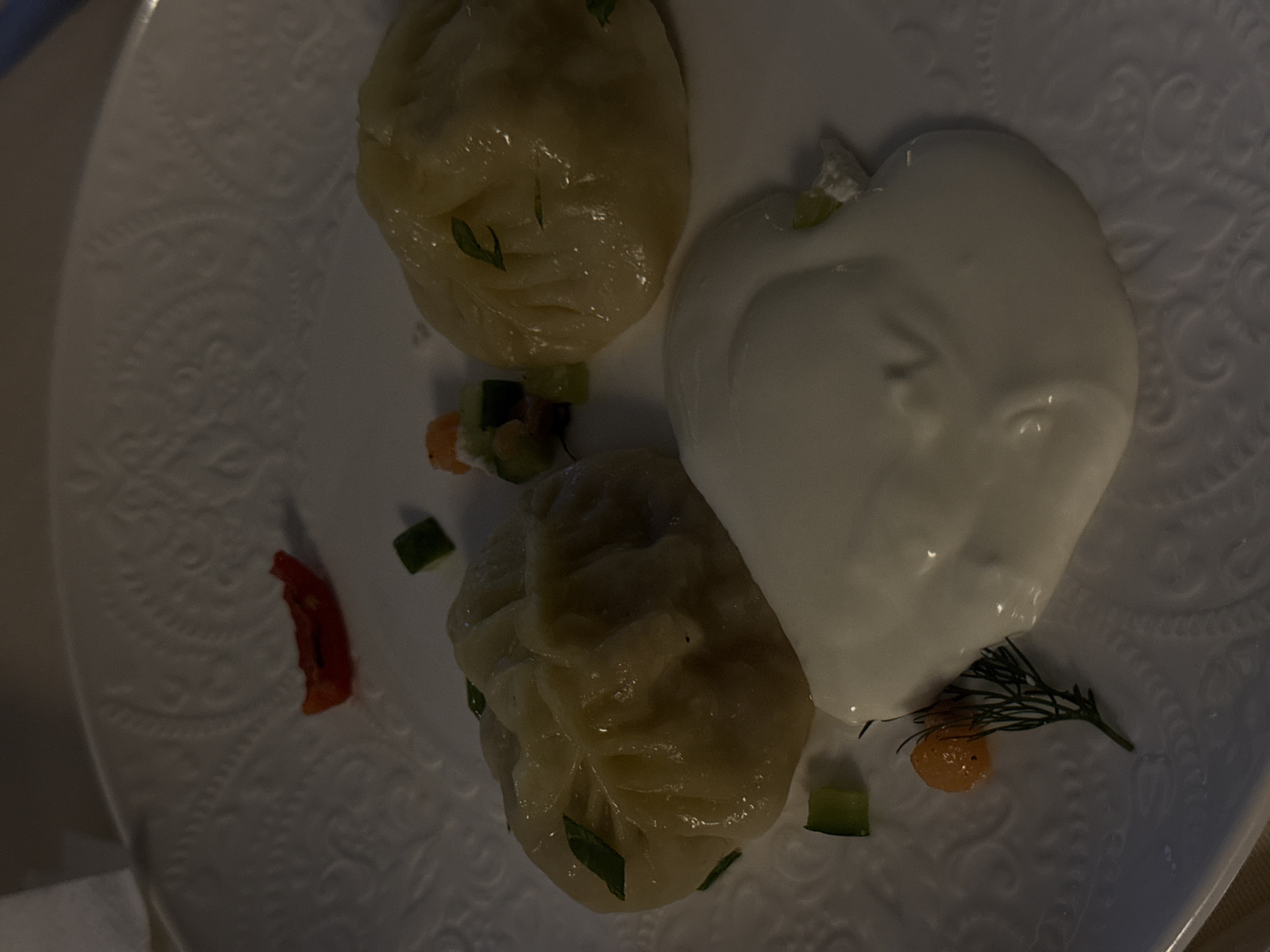 This image shows a plate of food, likely manti, a type of dumpling popular in Central Asia, including Kazakhstan. The dumplings are arranged on a decorative white plate. Two large, plump dumplings are visible, with a third partially obscured. They appear to be steamed and have a light, doughy exterior.
A dollop of a creamy white sauce, possibly yogurt or sour cream, is artfully placed next to the dumplings, shaped roughly like a heart. Scattered around the food are small pieces of chopped vegetables, likely cucumber and carrot, adding color and texture to the dish. A sprig of dill and a small orange garnish, possibly a tiny pepper or a cherry tomato, are also present. To the left of the dumplings, a small piece of red chili pepper is visible, suggesting a hint of spice.
The lighting in the image is dim, suggesting an indoor setting, possibly a restaurant or home dinner. The background is a textured white plate with an embossed pattern, adding to the presentation. The overall impression is of a well-presented and appetizing dish, typical of traditional Kazakh cuisine. There is no visible text in the image.