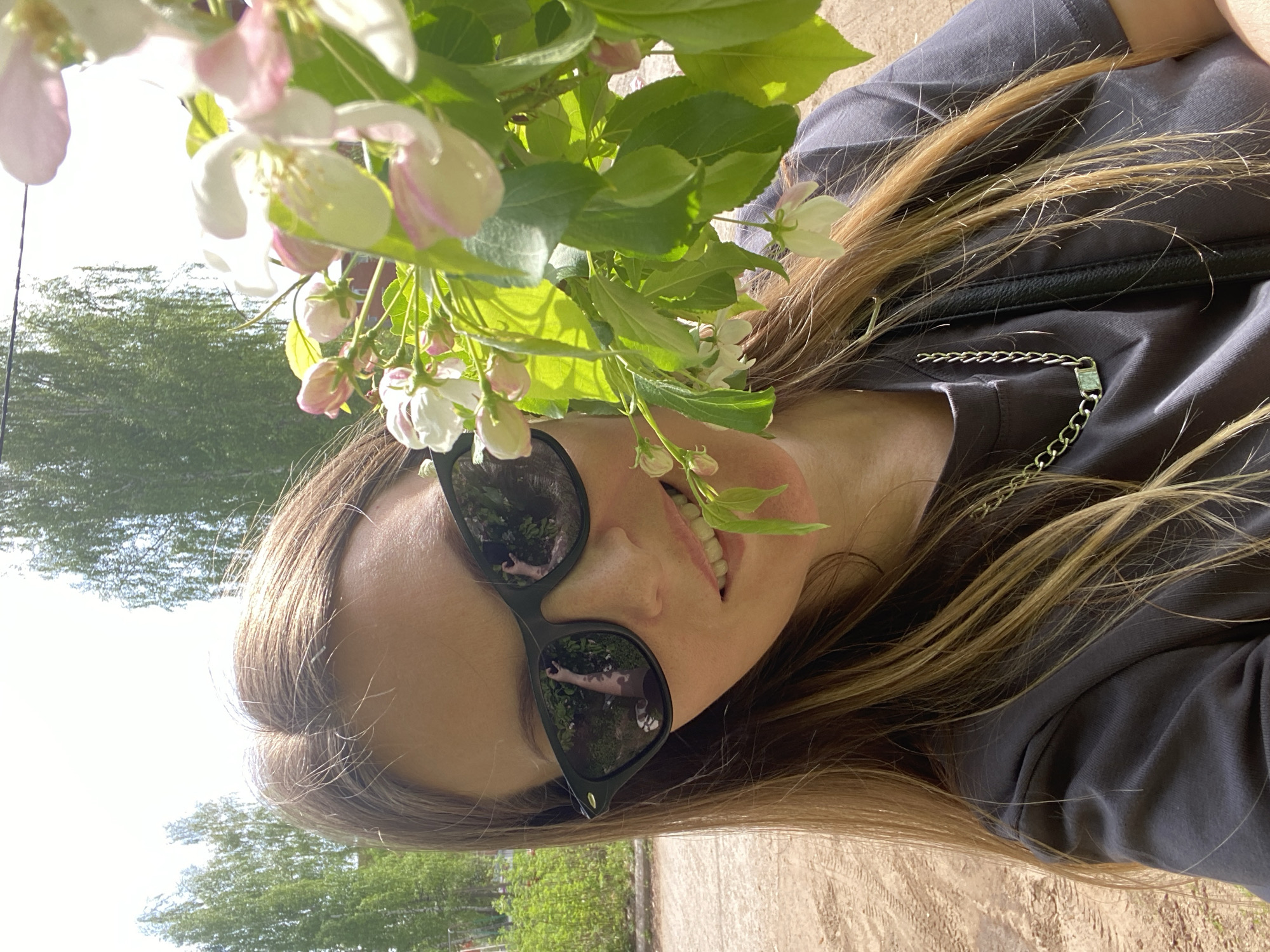 A young woman with long, light brown hair and sunglasses is holding a flowering branch with white and pink blossoms. She is wearing a gray t-shirt with a decorative silver chain. The sunglasses reflect an image of a person holding the phone. The background consists of trees and vegetation, suggesting a park or outdoor area. The scene appears to be taken in daylight. Based on the prompt, the location is Noviy, Russia.