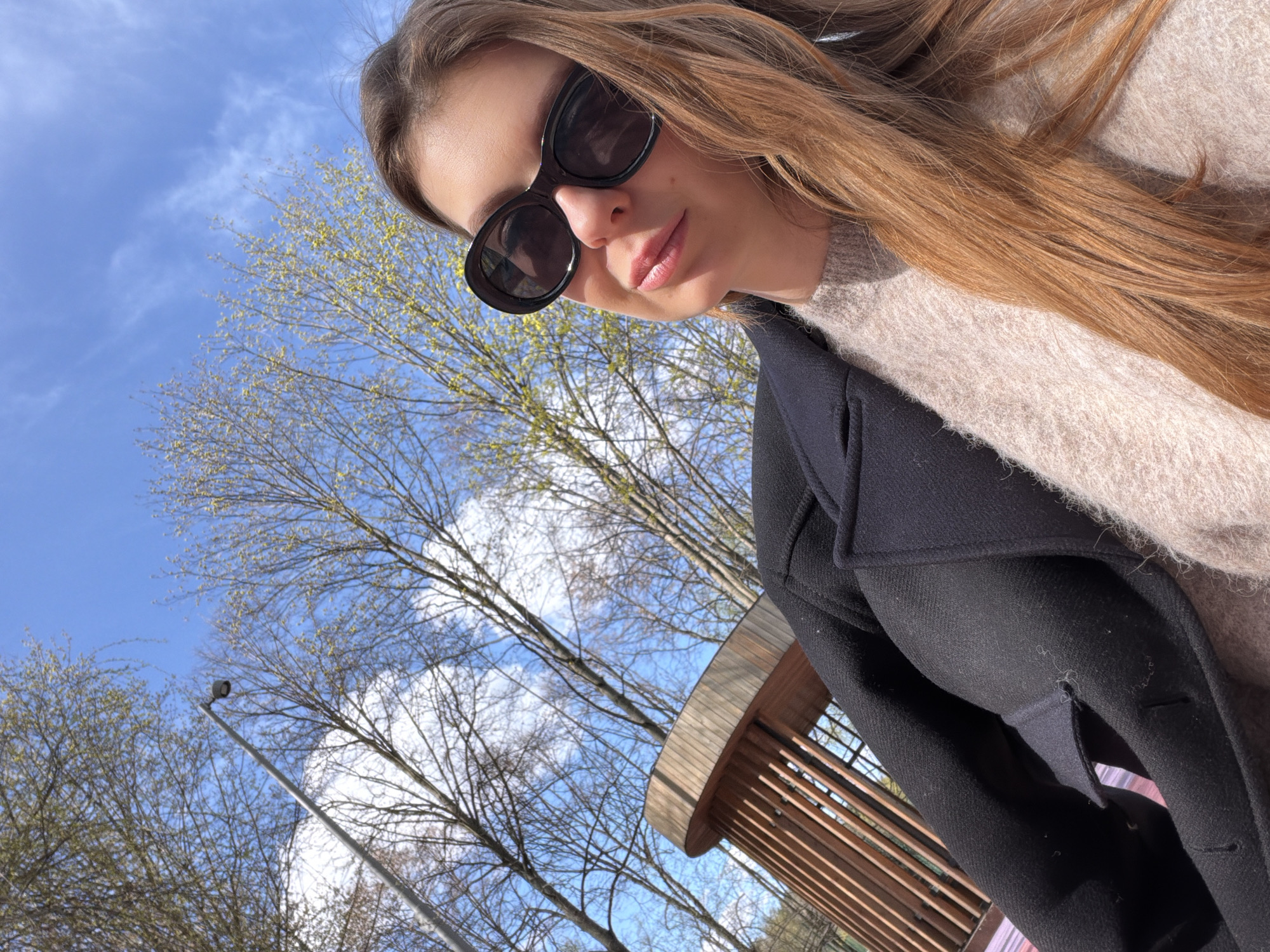 A woman wearing sunglasses, a light-colored sweater, and a dark coat poses outdoors. The background includes trees with budding leaves against a partly cloudy blue sky. A street lamp pole is visible among the trees. A wooden structure with vertical slats is positioned diagonally across the lower right portion of the frame. The photo was taken in Moscow, Russia.