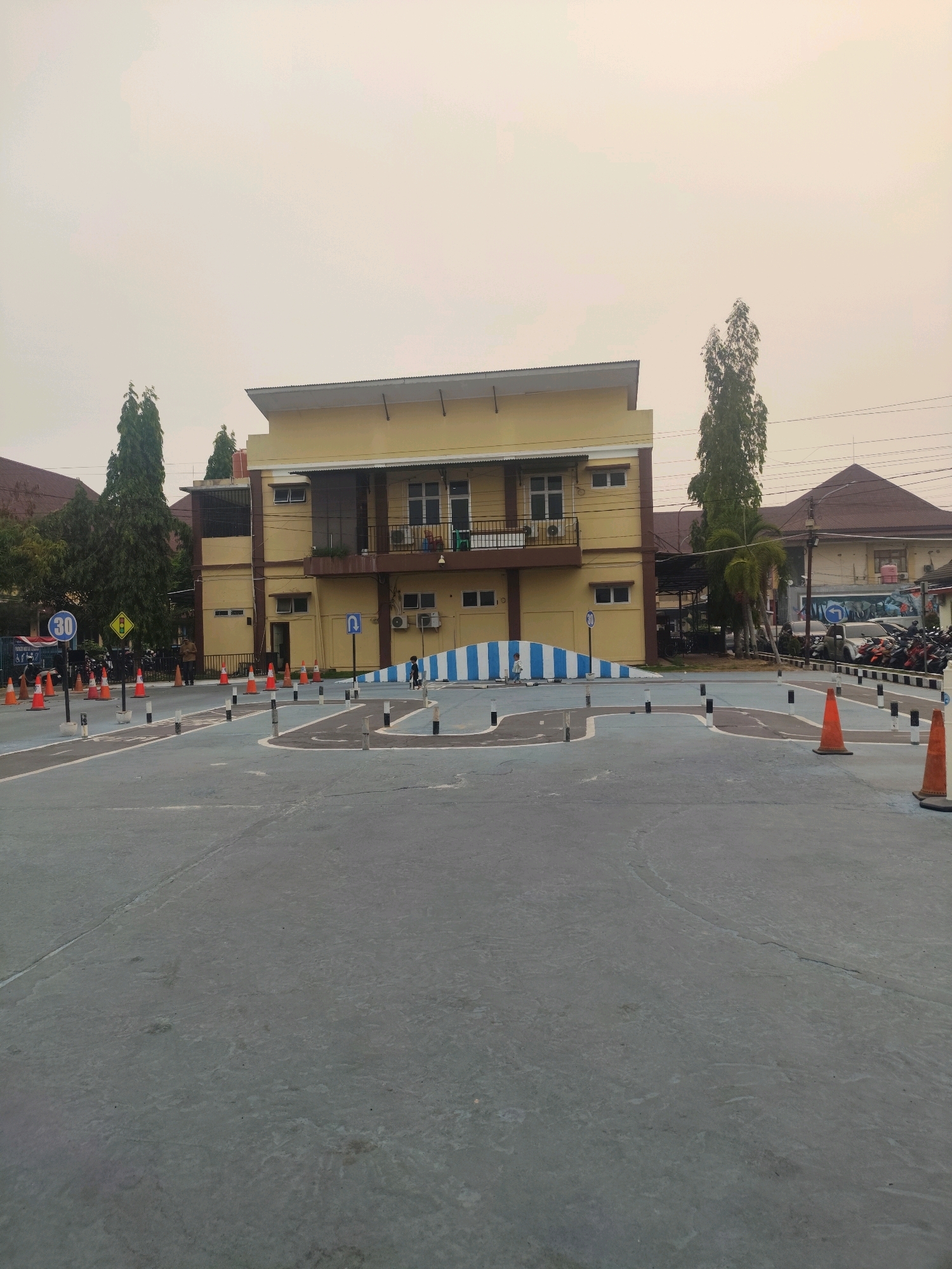 This is a ground-level, eye-level, wide-angle photograph of a driving practice track, likely located in Palembang, Indonesia. The track is made of grey asphalt with blue and white painted lines delineating the course. Orange traffic cones and black and white bollards are strategically placed to mark the course. In the background, a two-story yellow building with a dark-tiled roof stands prominently. The building has multiple windows and a balcony on the second floor. To the left of the yellow building, a dark-colored building with a tiled roof is partially visible. Several tall, slender trees are scattered throughout the scene. In the far background, a few motorcycles and cars are parked. The sky is overcast and a pale yellow color, suggesting either early morning or late afternoon light, or possibly haze. One person, appearing to be a child, is visible walking on the track in the middle ground. A blue traffic sign with a white circle and the number "30" inside is positioned to the left of the track. Another sign with a yellow diamond shape and black symbols is also visible near the "30" sign.
