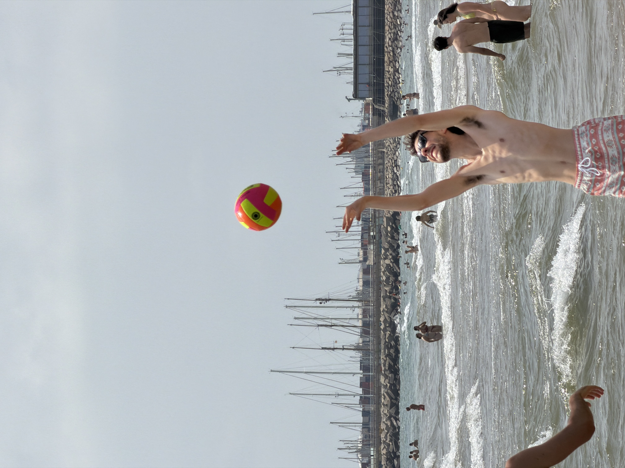 A man is playing volleyball on a beach in front of a breakwater. The man is in the foreground and is in the process of serving a ball. He is wearing red and white patterned shorts. The ball is orange and green and is in the air above his head. In the background, there are several sailboats and a breakwater made of rocks. There are other people in the water and on the beach. The sky is overcast.