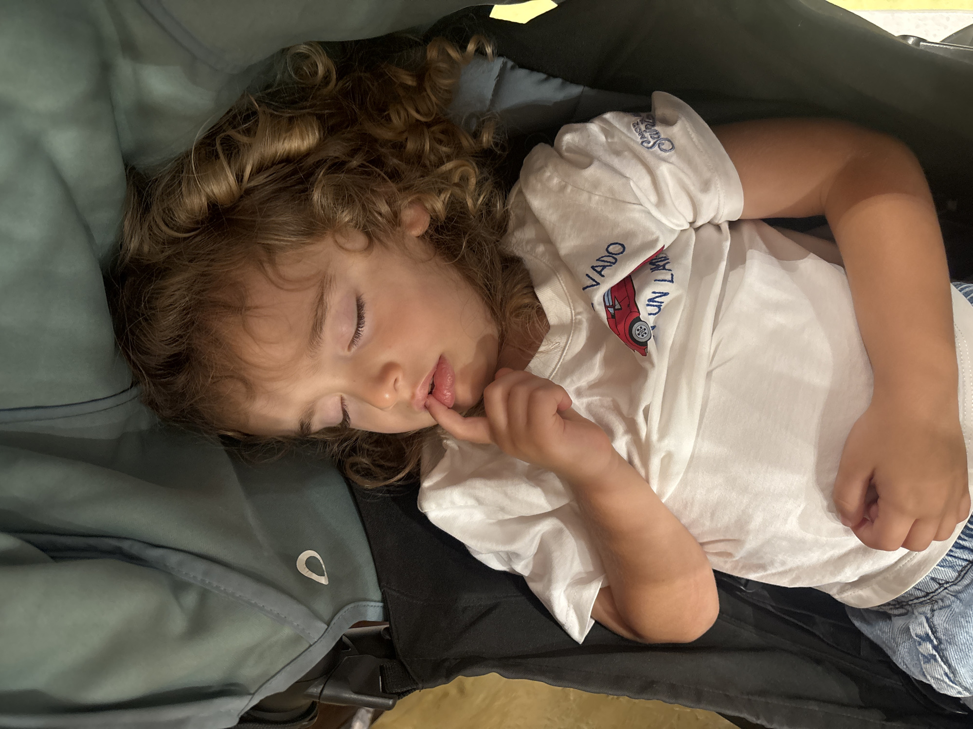 A young child with curly brown hair is sleeping, lying on their side. Their eyes are closed, and their finger is touching their lips. They are wearing a white t-shirt with blue lettering and a graphic of a red car. The child is nestled in what appears to be a stroller or carrier, with a soft, teal-colored fabric covering them on the left side. The background is dark, with a hint of yellow at the top. The image conveys a sense of peace and rest, suggesting a quiet moment during travel or an outing. The lighting suggests it could be daytime, but indoors or in shaded conditions. There is visible text on the t-shirt that reads "VADO UN LA" and another partially visible word above it.