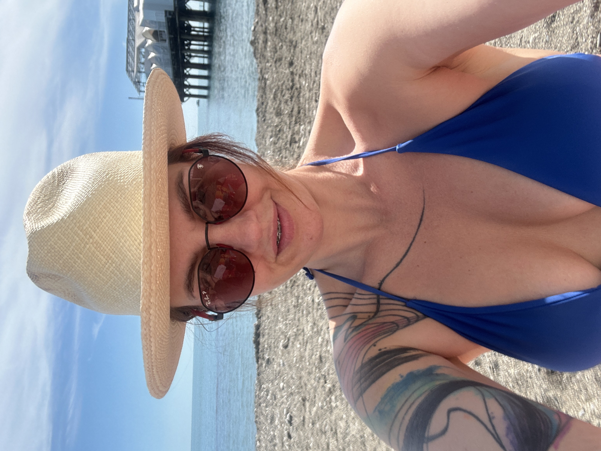 A cheerful woman is captured in a close-up, horizontal portrait, enjoying a sunny day at the beach in Sochi, Russia. She is smiling broadly, revealing braces on her upper teeth, and wears a vibrant blue bikini top. Her face is framed by a light-colored straw hat and large, round, brown-tinted sunglasses, on which the text "Ray-Ban P" is faintly visible. Visible tattoos adorn her left arm and extend onto her shoulder and collarbone, featuring abstract designs with various colors.
The setting is a pebbly or sandy beach, with the calm blue sea stretching into the background. A large, multi-storied pier or coastal structure, possibly a port facility or a complex building, is prominently visible over the water. The sky above is clear and bright blue, suggesting fair weather and strong daylight, likely mid-day or early afternoon. The woman appears relaxed and happy, posing for the photo in a leisurely seaside environment.