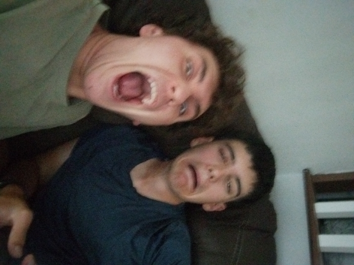 This indoor image shows two young adult males posing for a photograph, likely a selfie, within a residential setting. The individual on the left, positioned closer to the camera, has light brown, wavy hair and is wearing a light olive-green crew neck t-shirt. His mouth is wide open in an exaggerated expression, and his eyes are wide. The individual on the right, seated slightly behind and to the side, has short, dark hair and is wearing a dark blue crew neck t-shirt. His eyes are squinted, and his mouth is pulled downward in an exaggerated grimace. Both individuals are looking towards the camera. They appear to be seated on or against a dark brown upholstered couch or armchair, which occupies the mid-background. In the upper right background, a light-colored wall is visible, along with a partial view of what appears to be a white wooden balustrade or railing and a dark horizontal element, suggesting an interior space such as a living room. A hand is partially visible in the bottom right corner, possibly holding the device capturing the image. The precise location within Saint-Jean-sur-Richelieu, Canada, cannot be determined from the visual information provided in the image.