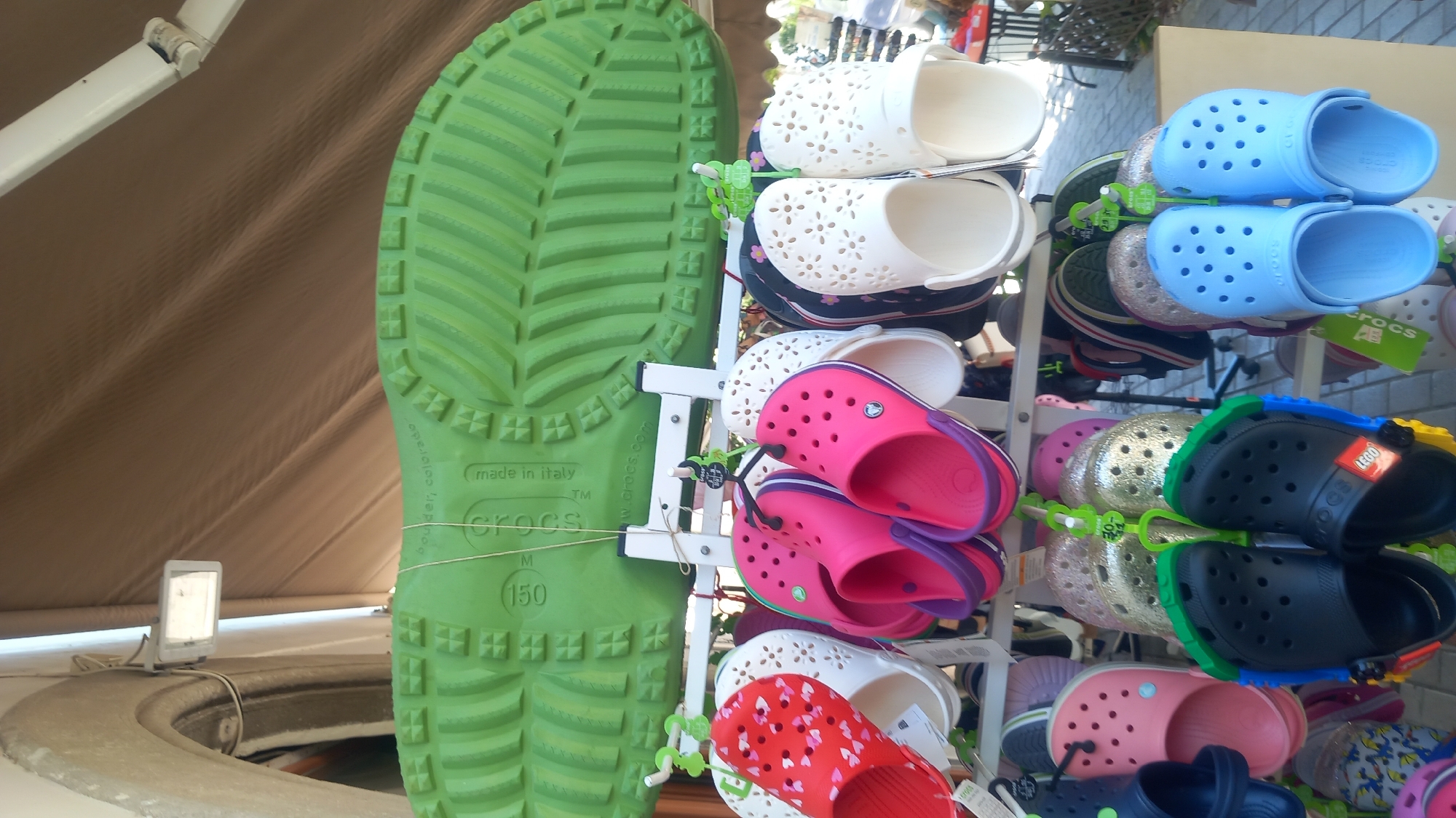 The image shows a display of Crocs shoes at a market stall in Kos, Greece. The shoes are hanging from metal racks, with a large green Crocs shoe sole displayed above them. The shoes are in various colors and styles, including pink, white, red with heart patterns, and black with LEGO accents. The background shows a tented area, suggesting an outdoor market. The lighting is natural, and the image is taken from a slightly low angle, looking up at the display.