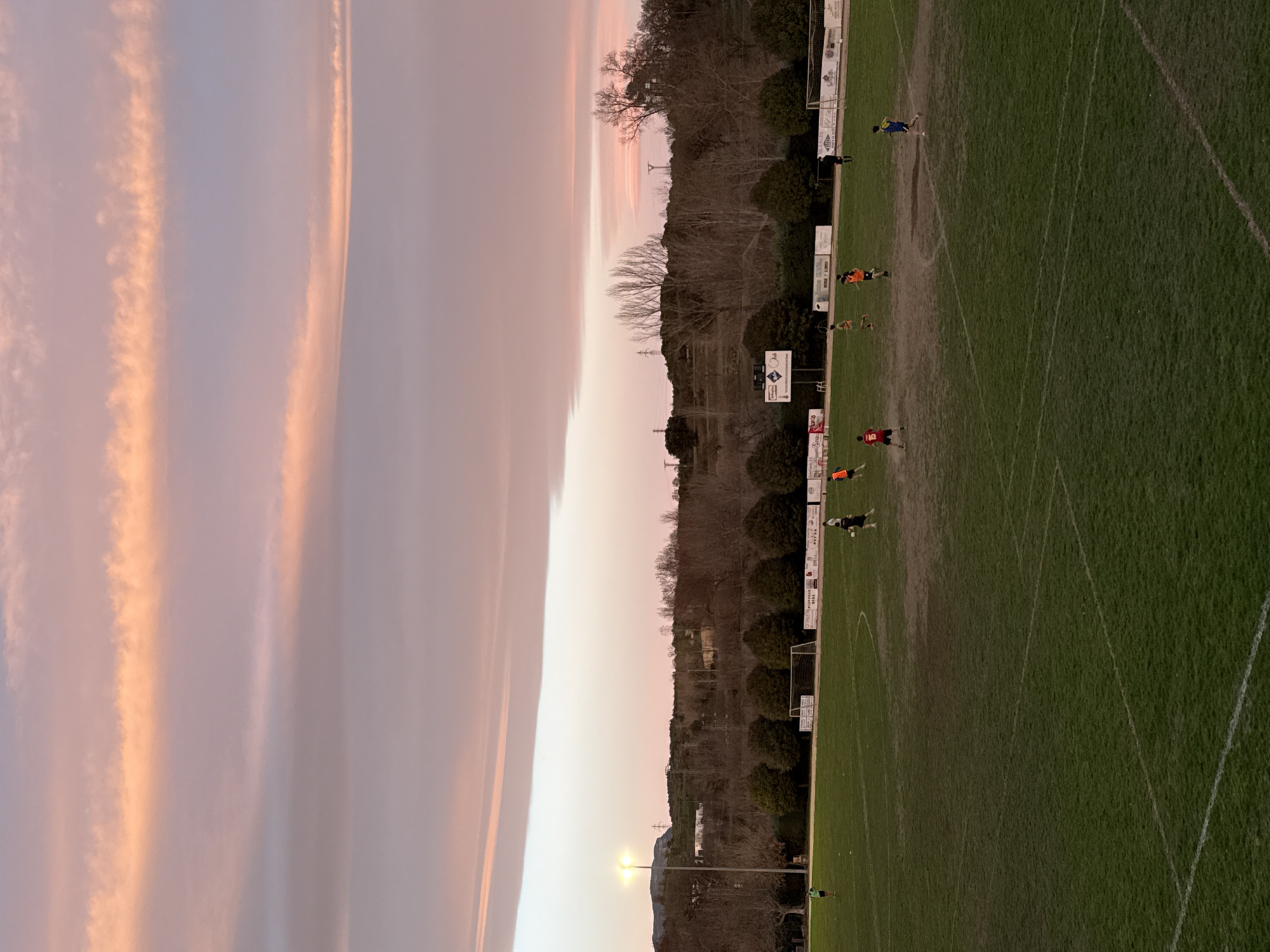 The scene depicts an outdoor football (soccer) pitch in Graus, Spain, captured during twilight or dusk. The upper two-thirds of the image show a sky with prominent, elongated clouds displaying striated patterns of purplish-pink, orange-pink, and pale blue.
The lower third of the image features a green grass playing field, marked with white lines indicating a football pitch, though some patches of ground appear worn. Ten individuals are present on the field. Eight individuals, appearing to be players, are scattered across the midground in various states of movement, suggesting a game or practice session. They are dressed in different colored jerseys, including orange, green, blue, red (one with the number "15" on the back), and white. One individual in a black top stands near the right-side goal. Two white football goals are visible at the far ends of the field, positioned near a hedge row and tree line.
A tall light pole equipped with two active luminaires casts artificial light onto the left side of the field. The background beyond the pitch consists of a dense line of mostly bare deciduous trees, followed by an undulating landscape of hills with visible utility poles and power lines. Advertising banners are affixed to the perimeter fencing behind the goals.