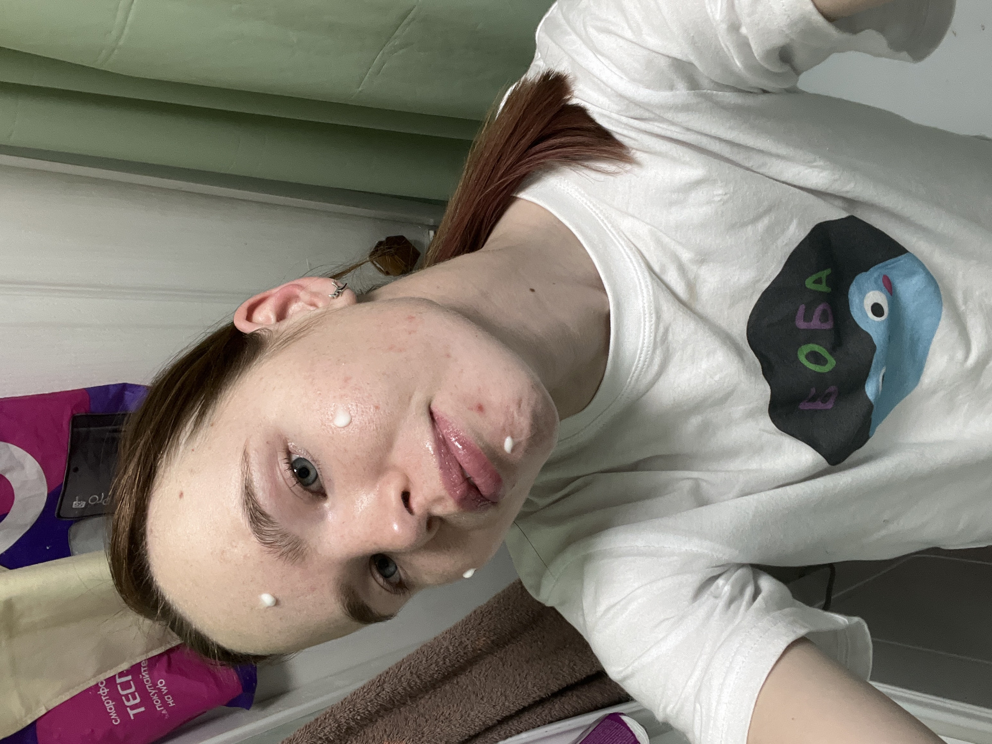 A young person with fair skin and light blue eyes is pictured from a low angle, with their head tilted slightly to the left. They have light brown hair with some reddish highlights, and a small silver earring is visible in their left ear. Four small dots of white cream are applied to their face: one on their forehead, one near their left eyebrow, one on their left cheek, and one next to their lower lip. They are wearing a white T-shirt with a design that includes the word "OBA" and a cartoonish blue figure.
The background is somewhat cluttered. To the left of the person's face, there is a pink package with Russian text. A brown towel and parts of other items are also visible. The photo appears to have been taken indoors, possibly in a bathroom.
There is no information in the image to indicate this was taken in Chaikovsky, Russia.