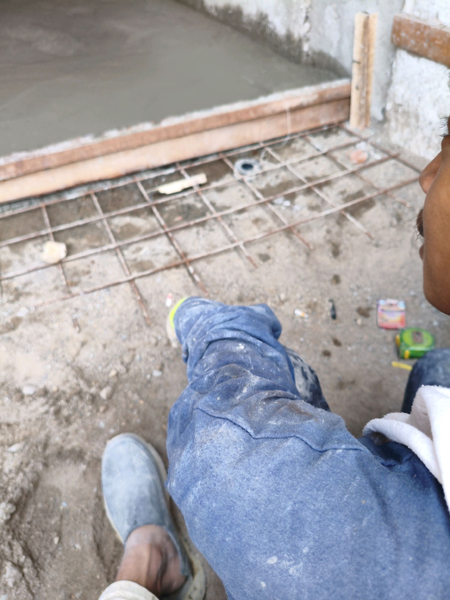 This image depicts a construction scene, possibly in the city of Bekasi, Indonesia, focusing on the legs and feet of a person wearing blue denim work pants and grey work shoes. The person is seated or resting. In the foreground, loose gravel and dirt cover the ground. Behind the person, a section of freshly poured concrete is visible, separated by wooden framing and a grid of rebar. A small, green tape measure and a pink and orange object, possibly a small box or packaging, are on the ground near the rebar. The overall impression is one of ongoing construction work.