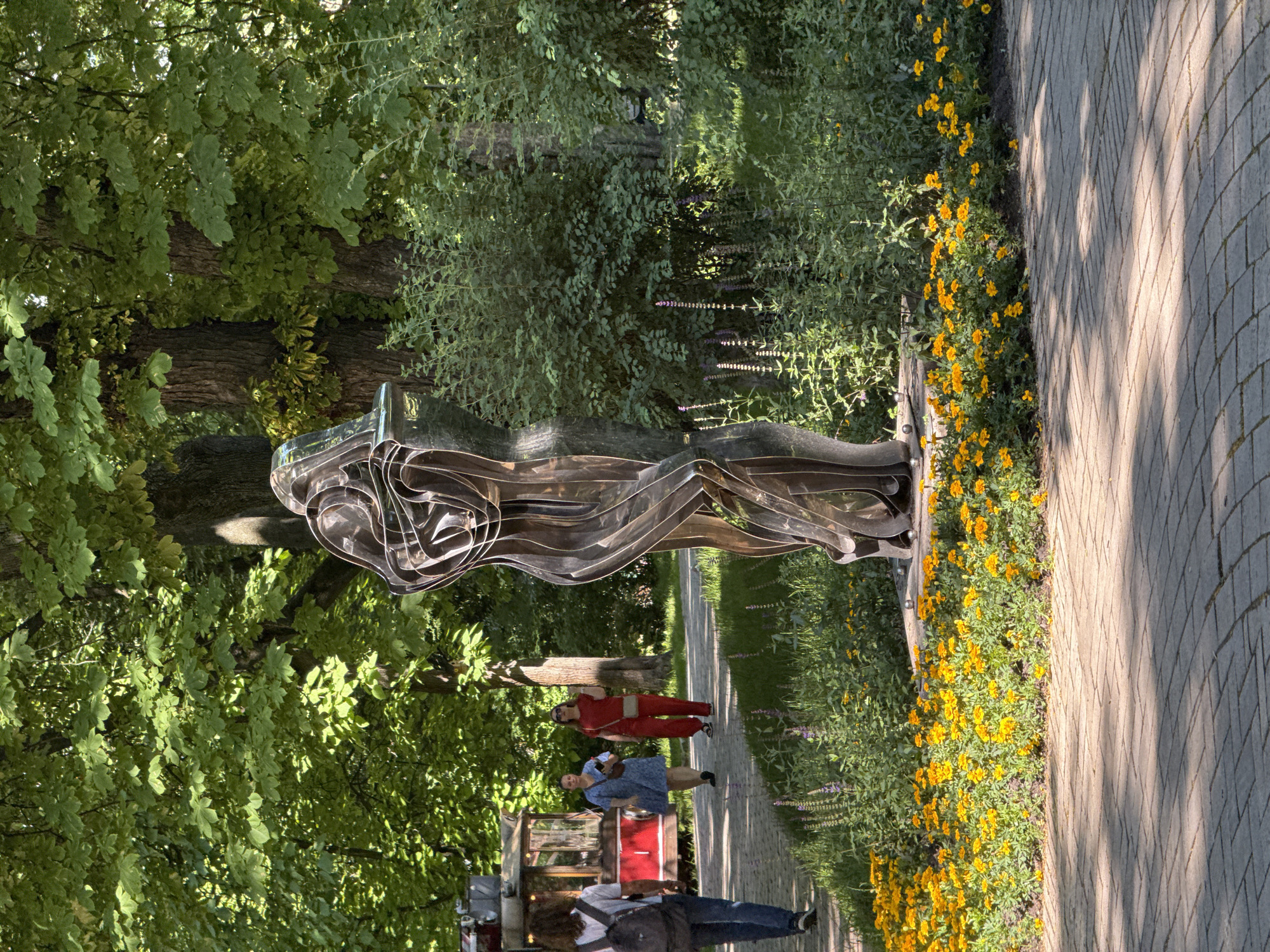 In a sunlit park, a modern abstract metal sculpture, depicting entwined figures, stands prominently. In the background, a red food truck is visible, and two women are walking on a paved path. One woman is wearing a blue dress and carrying a purse, while the other is dressed in a red jumpsuit and also carries a purse. A person with a backpack is walking away from the camera on the same path. The scene is bordered by a vibrant bed of yellow marigolds and lush green foliage, with large trees providing a canopy overhead. The paved path is laid with grey interlocking bricks, dappled with sunlight and shadows. This scene is in Kyiv, Ukraine.