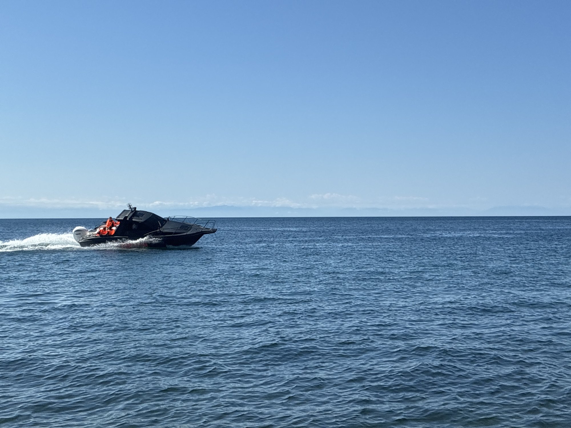 A black speedboat with two people wearing orange life vests is moving quickly through the blue water of Lake Baikal, kicking up a white wake. The boat is in the left half of the frame, with the vast expanse of the lake and a clear blue sky filling the rest of the image. Distant, hazy mountains can be seen on the horizon. The image appears to have been taken from a stationary boat or the shore, with the speedboat moving away from the camera.