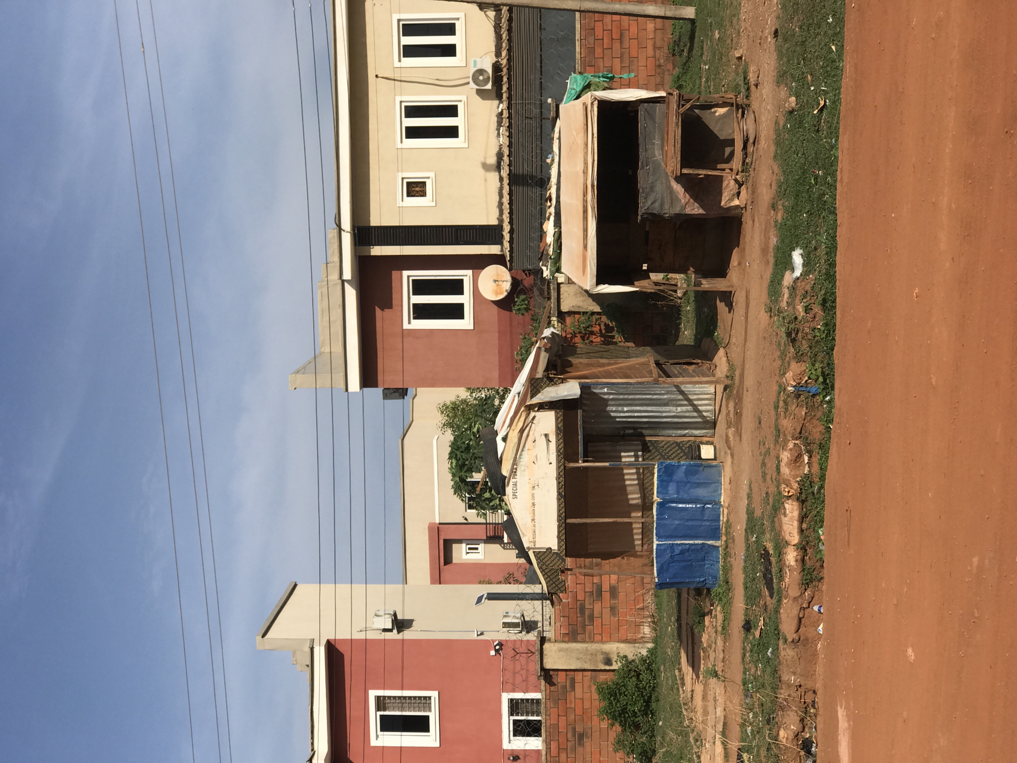 The image shows a street scene in Abuja, Nigeria. A row of multi-story buildings in varying shades of red and beige is visible on the left side of the frame, with visible windows and electrical wiring running along the front. Adjacent to the buildings is a dirt path alongside the street, which has several informal stalls or small shops constructed with corrugated metal and wood. Some of the stalls are covered with blue tarpaulins. The street is made of red dirt and extends along the right side of the image. The sky is light blue with a few scattered clouds.