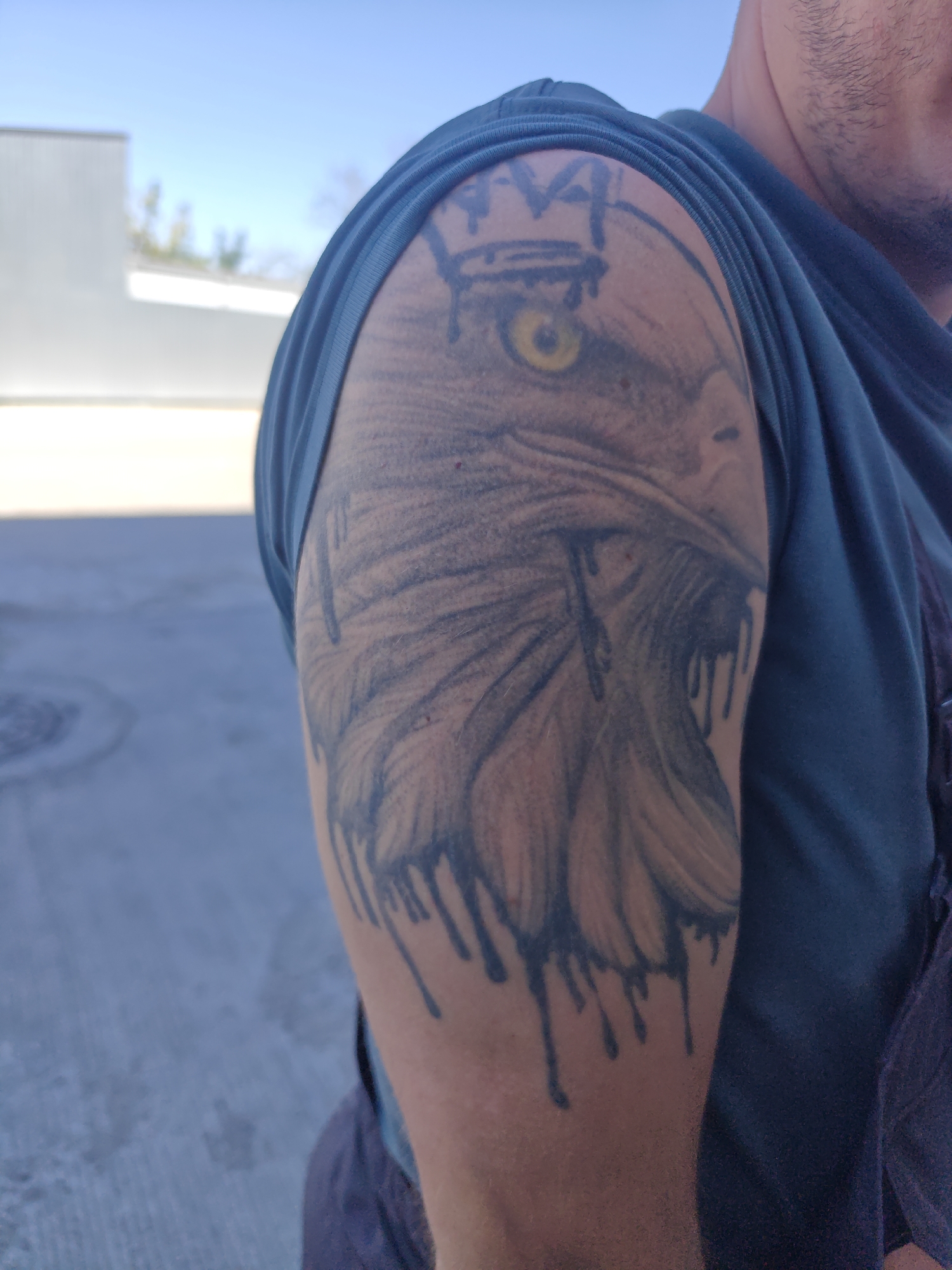An outdoor, medium shot captures a person's upper arm and shoulder, adorned with a dark, detailed tattoo of an eagle's head. The tattoo features a prominent yellow eye, a crown-like design above it, and a dripping effect below. The person is wearing a dark blue t-shirt. The background consists of a bright blue sky and a blurry, light-colored building with some greenery in the distance. The ground appears to be paved. The image does not provide any specific visual cues to indicate a location in Krasnodar, Russia.