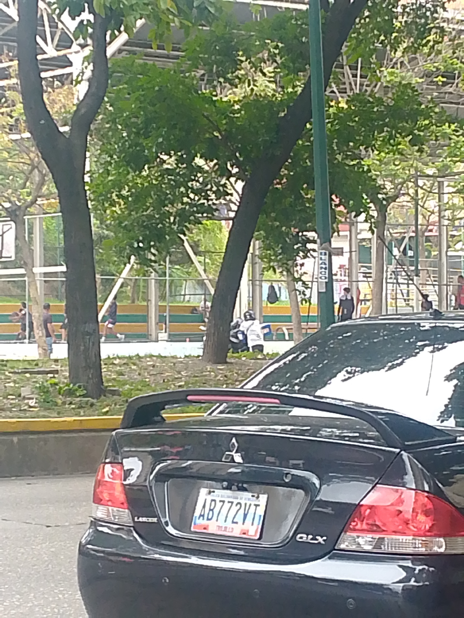 The image shows a black Mitsubishi Lancer GLX car with license plate AB772VT in Caracas, Venezuela. In the background, there is a basketball court with people playing basketball. The court is surrounded by trees and buildings. A street sign reading "Blanco" is visible near the court.