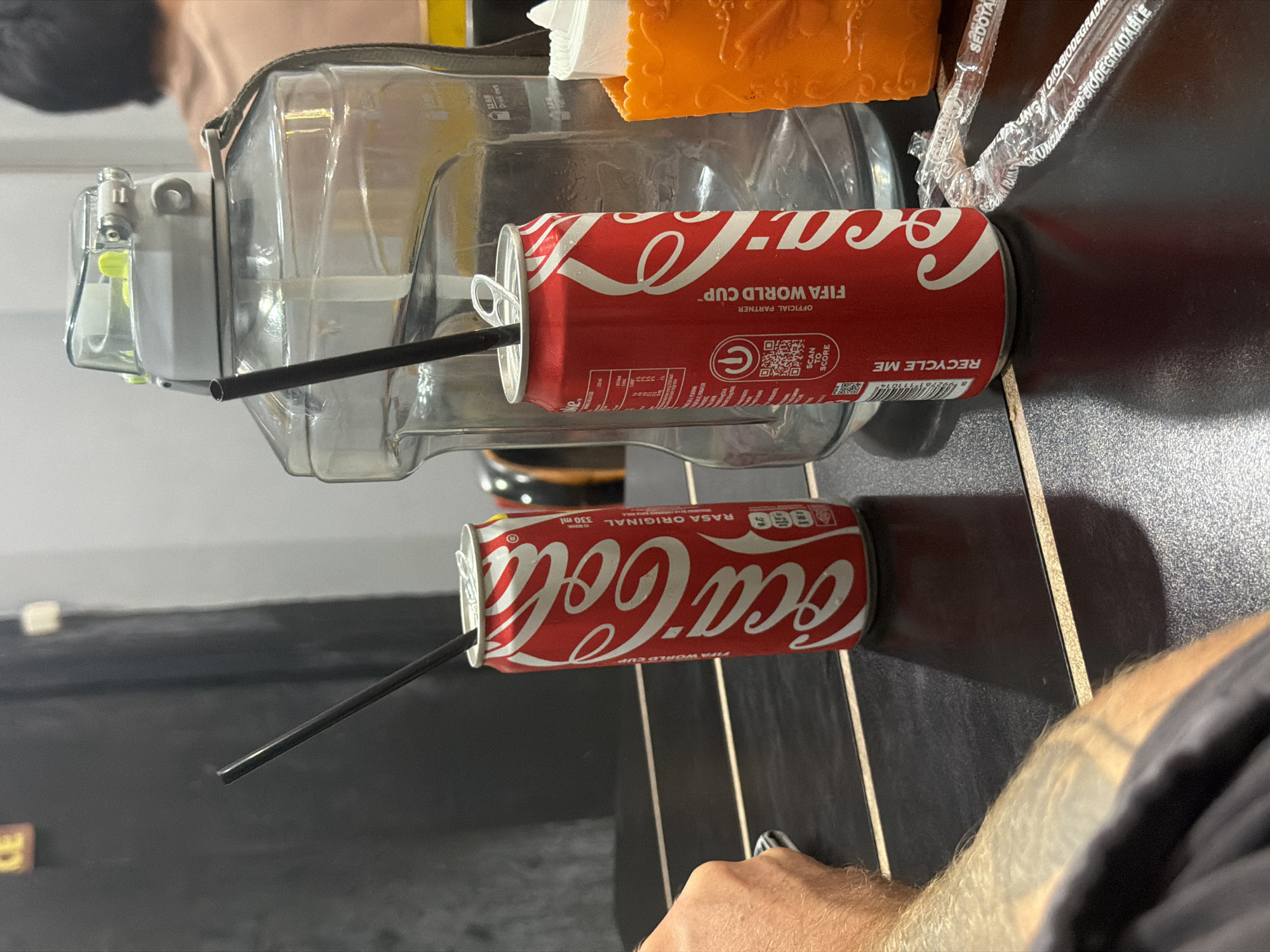 In Kuta, Indonesia, a black plastic table holds two open cans of Coca-Cola, each with a black straw inserted. To the right, a large clear plastic water jug with a gray lid and strap is positioned behind the cans. An orange plastic holder containing white rolled items, possibly napkins, is visible to the far right. The background shows a dark vertical surface and a light gray wall. A person's tattooed arm is in the foreground on the left.