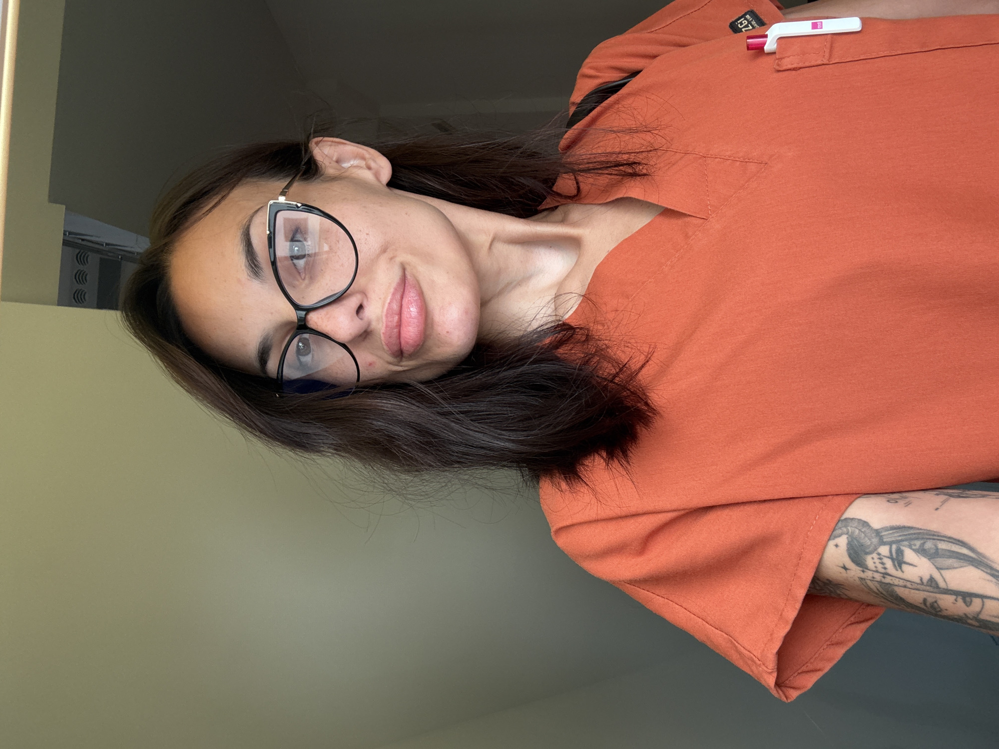 A selfie taken indoors shows a woman with dark hair and glasses. She is wearing an orange scrub top. She has tattoos on her left arm, which is visible. A pen is tucked into the pocket of her scrub top. The background appears to be a wall, with a vent visible at the top. The location is not specified.
