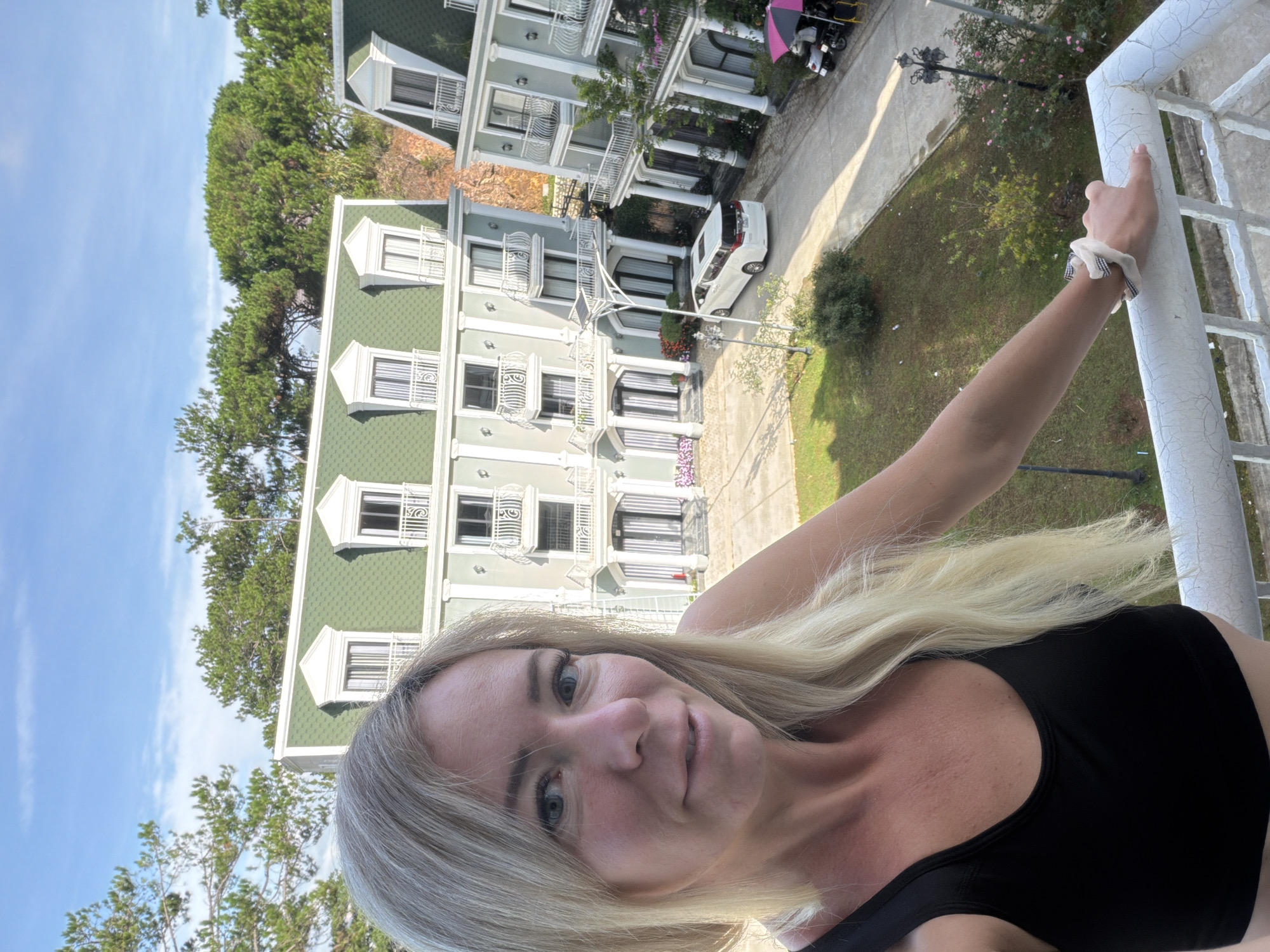 A woman with blonde hair is taking a selfie on a balcony, with a light green building and trees in the background. The building has a distinctive mansard roof with dormer windows and ornate balconies. A white minivan is parked in front of the building. The scene appears to be in a courtyard or street area with some landscaping. The woman is wearing a black tank top and has a scrunchie on her wrist. The sky is blue with some clouds, suggesting it is daytime. The location is identified as Xuân Trường - Đà Lạt, Vietnam.