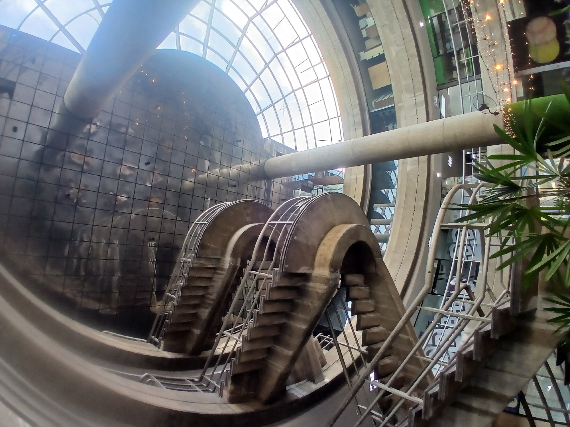 The image shows the interior of the headquarters of the state-owned company CVG Ferrominera Orinoco in Ciudad Guayana, Venezuela. The building's architecture features circular stairs with metal railings and a large, curved, glass-paneled wall. A wide cylindrical pipe runs horizontally across the upper section of the frame. Palm fronds are visible in the foreground, partially obscuring the structures.