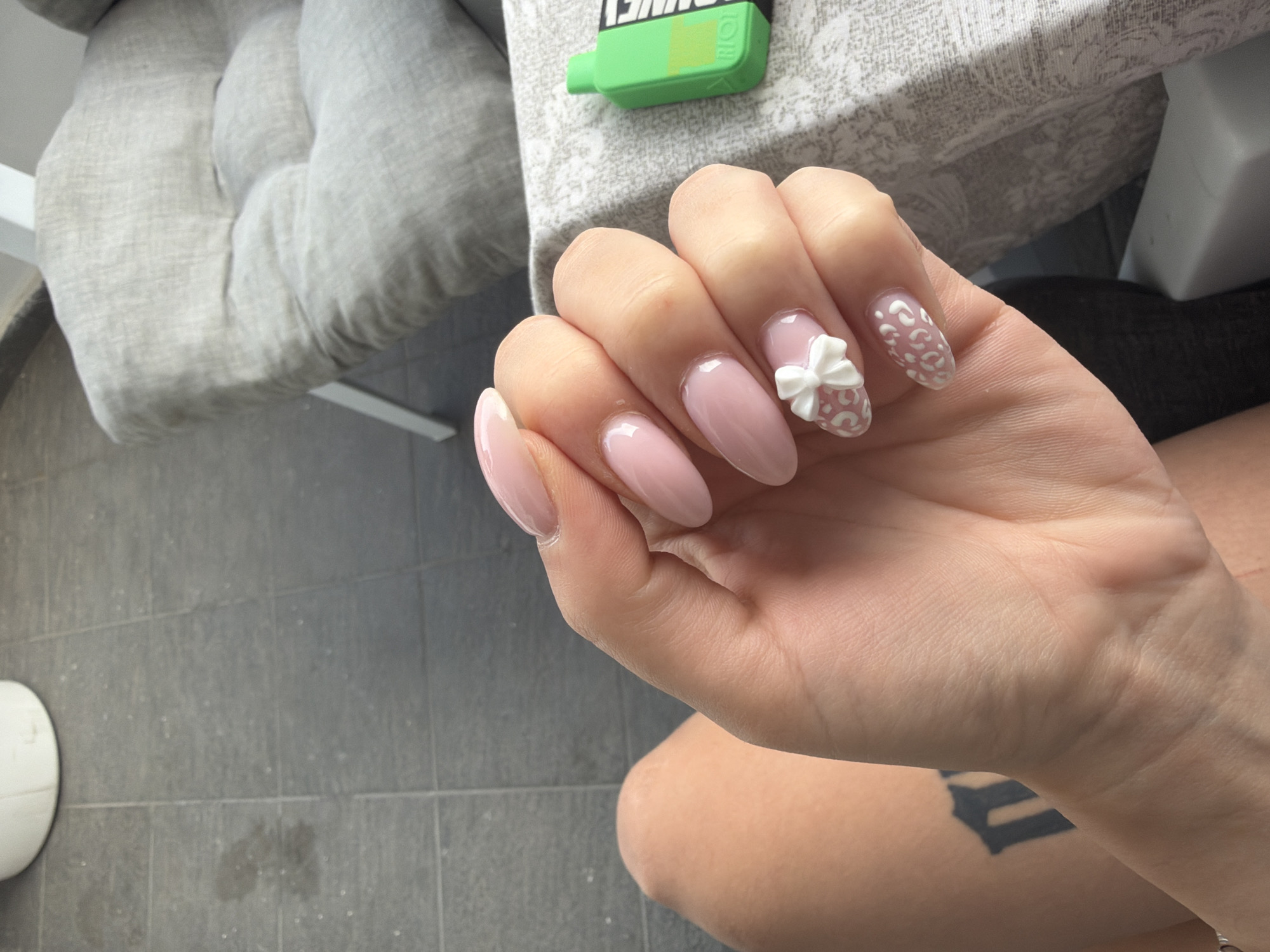 The image shows a close-up of a person's hand with light pink almond-shaped nails. Three nails are visible: the index finger, middle finger, and ring finger. The index and middle fingers have plain light pink polish. The ring finger has a white 3D bow on it, and the little finger appears to have a similar white bow. A tattoo is visible on the person's forearm. The background includes a grey tiled floor, a light-colored cushioned chair, and a bright green object, possibly a vape pen, resting on a patterned fabric. The setting appears to be outdoors, possibly a patio or balcony. There is no information to determine the location in Plovdiv, Bulgaria.