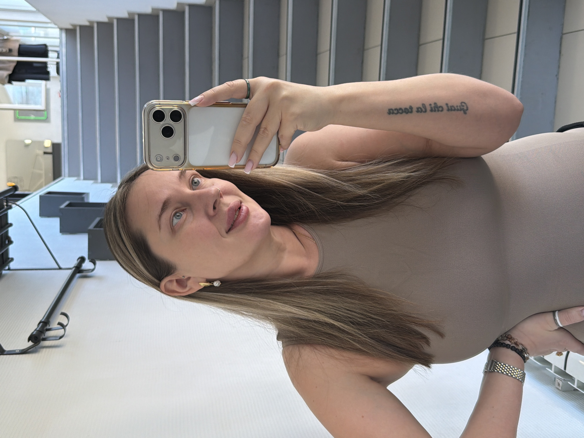 A woman is taking a selfie with her phone in what appears to be a modern interior, possibly a building in Chaikovsky, Russia. She has blonde hair with darker roots and is wearing a neutral-colored top. Her left arm is visible, adorned with multiple bracelets, and a tattoo on her right arm is partially visible. She is wearing a ring on her left hand. The background features a white wall with vertical lines, resembling a modern staircase design. A garment rack and mirror are visible in the background. The overall lighting is bright and even, suggesting indoor lighting.