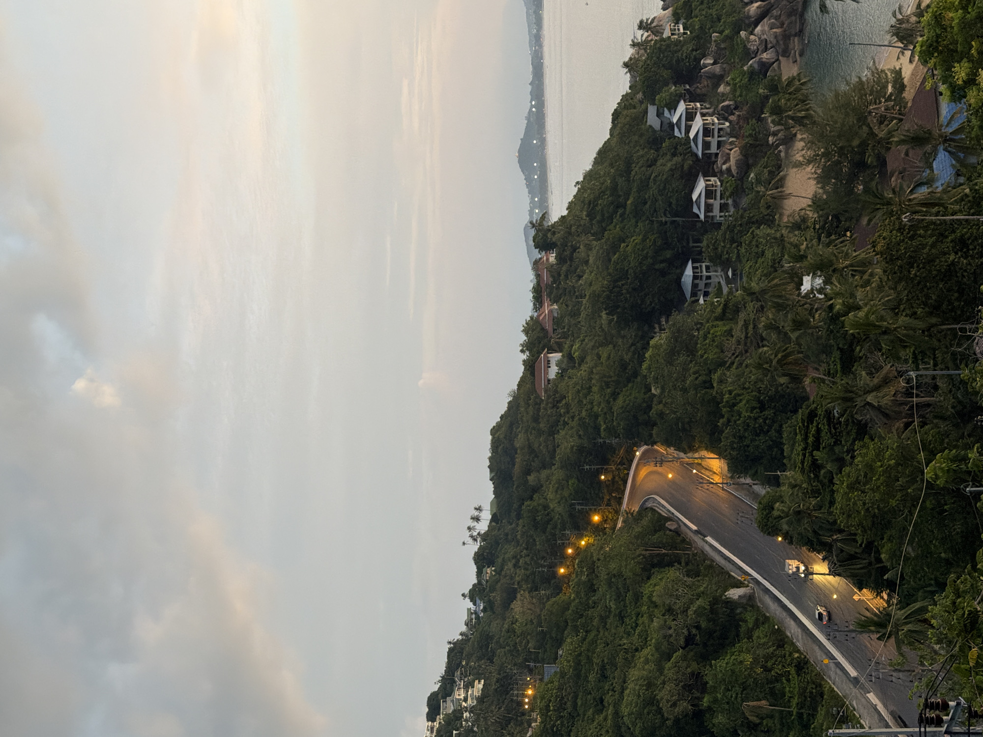 A scenic overlook captures a winding road ascending a lush, tropical hillside on the island of Ko Samui, Thailand, with the ocean stretching out in the distance. The road, illuminated by warm streetlights, curves through dense green foliage and is bordered by electric poles. Several vehicles, including a white van and two cars, are visible on the road. On the hillside, white houses with tiled roofs are scattered amongst the trees, some appearing to be part of a resort or residential development. A small sandy beach is visible near the water's edge, bordered by rocks and palm trees. The sky above is overcast with soft, diffused light, suggesting either dawn or dusk. In the far distance, other islands or peninsulas are faintly visible on the horizon, with scattered lights indicating human habitation. The overall impression is one of serene natural beauty and coastal development.