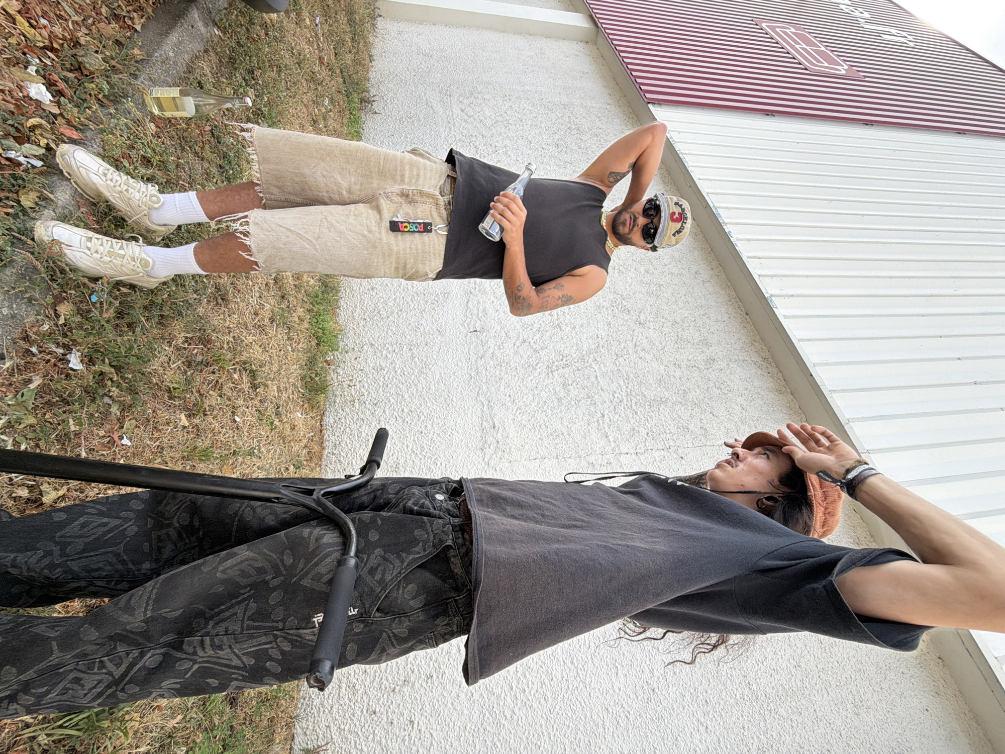 The image shows two individuals standing outdoors. The person on the left, wearing a black tank top, tan shorts, and a baseball cap, holds a clear bottle. The person on the right, in a dark hoodie and patterned pants, stands next to a scooter. They are positioned in front of a building with white and red vertical siding. The ground is covered in dry grass and fallen leaves. The scene appears to be in an urban or suburban setting. Based on the style of the building and the presence of urban elements, this location could plausibly be Caluire-et-Cuire, France, a commune on the outskirts of Lyon.