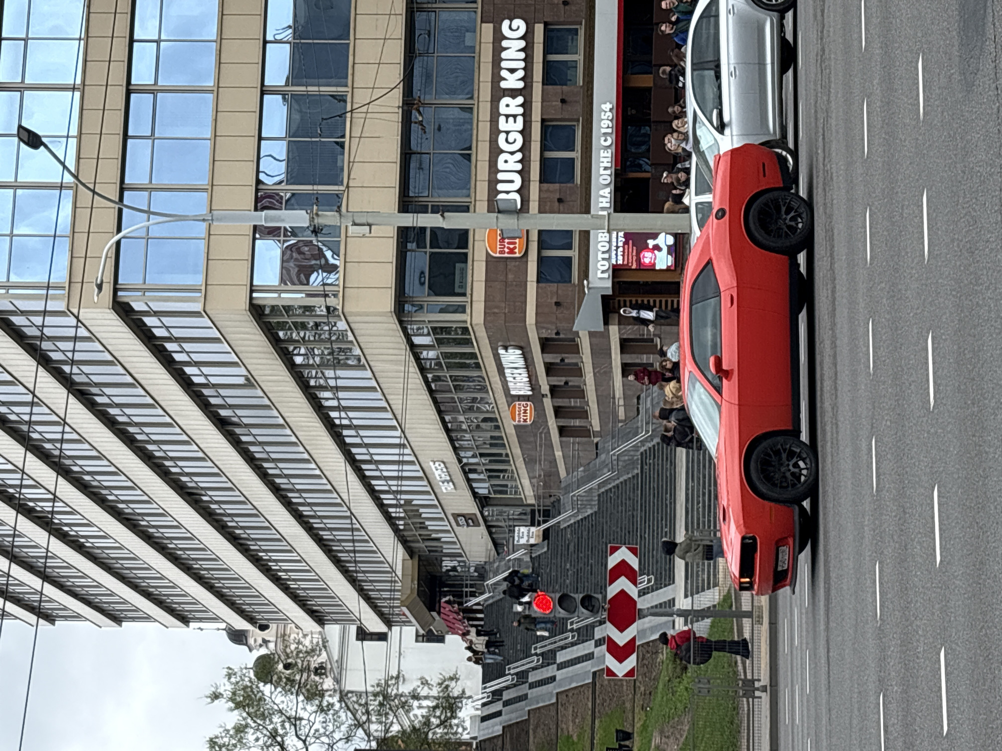 Here's a detailed description of the image:
**Location:** The image appears to be taken in Minsk, Belarus, based on the Cyrillic text visible on the Burger King signage ("ГОТОВІ НА ОГНЕ С 1954").
**Scene Description:** The image depicts a street scene with a multi-story building on the left and a road on the right. The building houses a Burger King restaurant. A red car is parked on the right side of the road. People are standing on the stairs leading up to the restaurant and on the sidewalk. A traffic light is visible, showing a red light. A street lamp post is on the left. A white car is parked behind the red car.
**People:** There are several people visible on the stairs and sidewalk in front of the building. Some appear to be customers of Burger King, while others may be pedestrians.
**Objects:** Key objects in the image include the multi-story building, Burger King signage, the red car, the white car, the road with lane markings, the traffic light, the street lamp post, stairs, and various architectural elements of the building.
**Environment:** The environment appears to be an urban setting with modern buildings and infrastructure. The weather seems overcast, as indicated by the lighting and cloud cover.