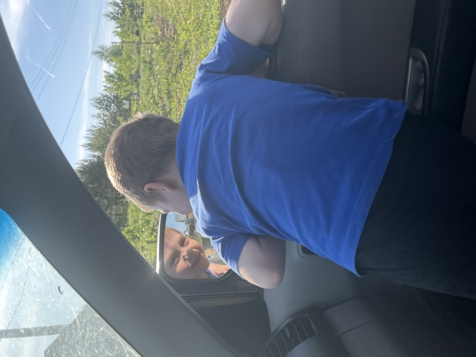 A boy in a blue shirt and black pants peers into the side mirror of a gray vehicle, his reflection visible in the mirror. The background shows a field with trees and a blue sky with contrails. The location appears to be outside, as indicated by the natural light and landscape. The scene does not appear to be within the city limits of Фоки, Russia.