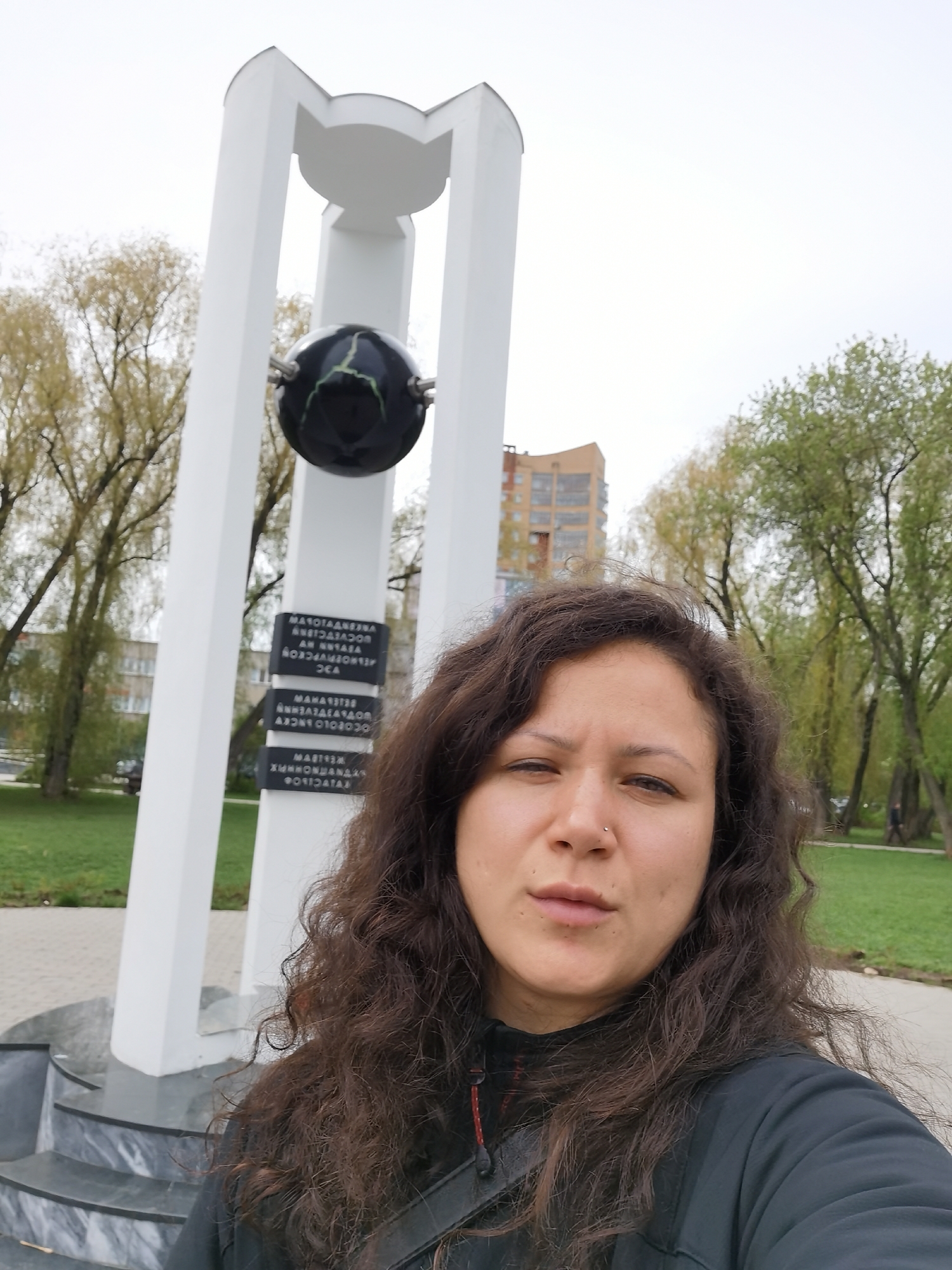 A selfie of a woman with dark, curly hair and a nose piercing, taken outdoors near a white monument featuring a black globe. The monument has four white columns supporting a disk-shaped top. Below the globe, there are plaques with text in Russian. The location is in the city of Perm, Russia. Behind the monument, there's a park with green grass and trees, and a multi-story brick building in the background. The sky is overcast. The woman is wearing a black jacket. The monument is situated on a platform with marble steps.