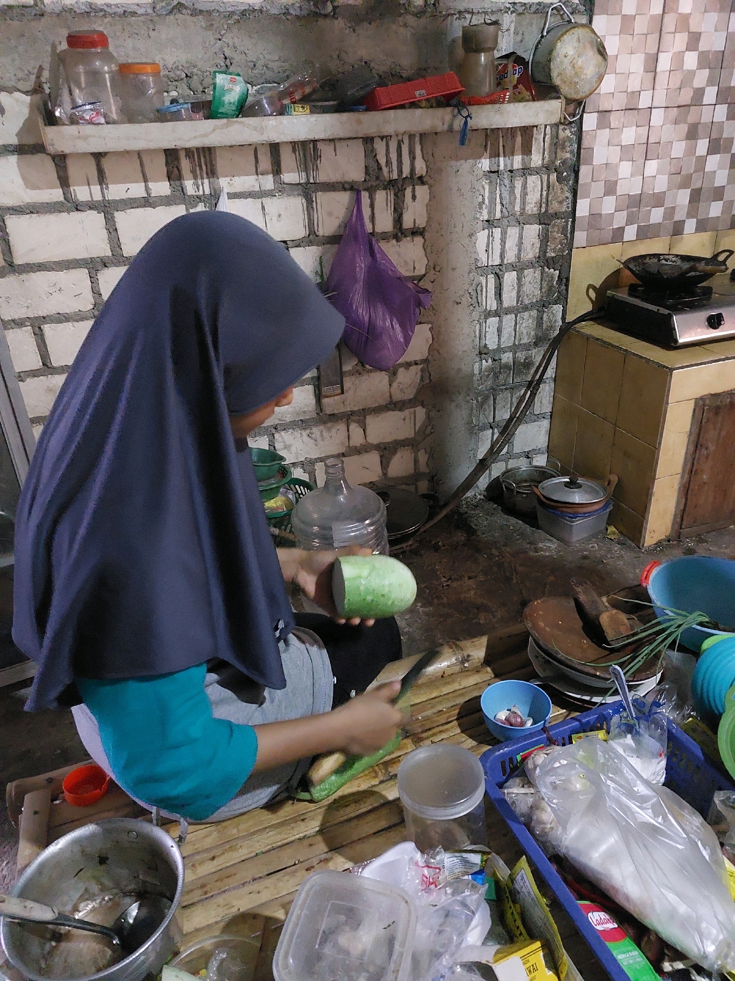 In Sarangan, Indonesia, a person wearing a dark blue hijab and a teal shirt is preparing food. They are seated on a bamboo mat in what appears to be an outdoor kitchen or a rustic indoor space with brick walls. The person is holding a cucumber in their left hand and a knife in their right, in the process of cutting it.
The environment is cluttered with various kitchen items. To the left of the person is a large metal pot containing remnants of food and a ladle. Various plastic containers, bowls, and bags of ingredients are scattered around. In the background, there is a gas stove with a pan on top, and shelves lined with jars and containers. A purple plastic bag hangs on the wall. The overall impression is one of a functional, albeit unkempt, cooking area.