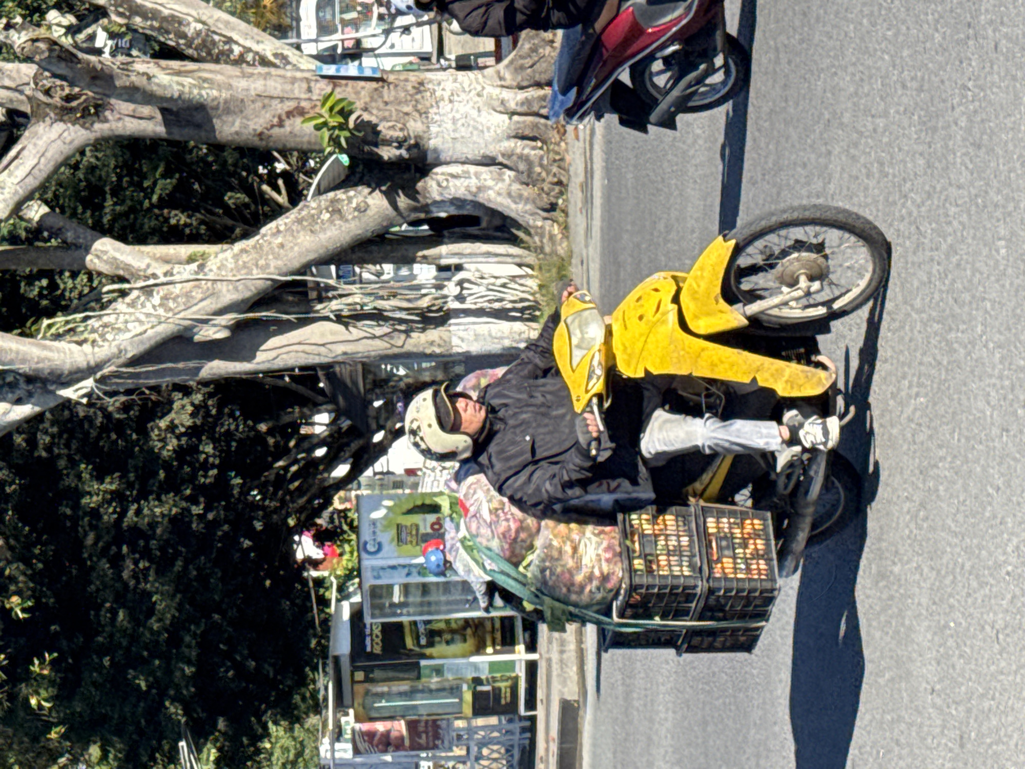 A man wearing a helmet and a dark jacket is riding a yellow scooter loaded with produce. The scooter has two black crates stacked on the back, filled with fruits and vegetables. The man is looking forward, and the scooter is in motion on an asphalt road. In the background, there are trees, a building with signage, and another scooter. The scene appears to be a street in Phường Xuân Hương, Đà Lạt, Vietnam, indicated by the style of the buildings and vegetation.