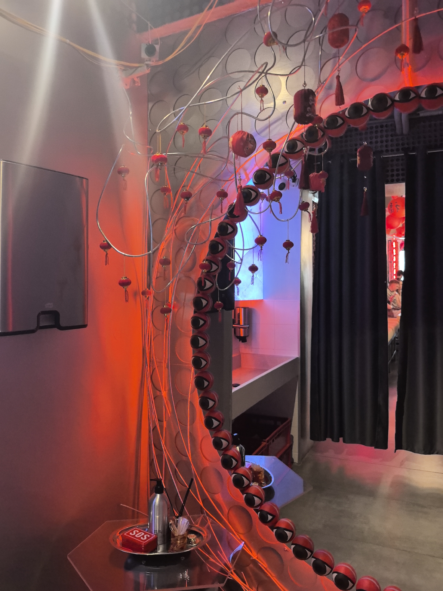 The image depicts the interior of what appears to be a restroom or wash area, possibly in a commercial establishment. The dominant color palette is red, due to intense artificial lighting. On the left, a silver hand sanitizer dispenser is mounted on a grey wall. In the foreground, a metallic tray holds a red "SOS" box, a pump bottle dispenser, and a glass container with thin sticks.
The central and right portions of the image are dominated by a large, red, eye-themed art installation. This installation consists of numerous red spheres with black and white eye motifs, suspended from thin metal wires that curve and intertwine. Small red lanterns with tassels are also hanging from the wires, adding to the decorative and somewhat whimsical nature of the piece.
Behind the installation, a bathroom sink and countertop are visible, illuminated by blue and pink lights. To the far right, black curtains obscure part of the background, but through a gap, a glimpse of another room is visible, showing people and red lanterns. The flooring appears to be concrete.
The image does not contain any discernible elements that would specifically indicate the location as Perm, Russia.