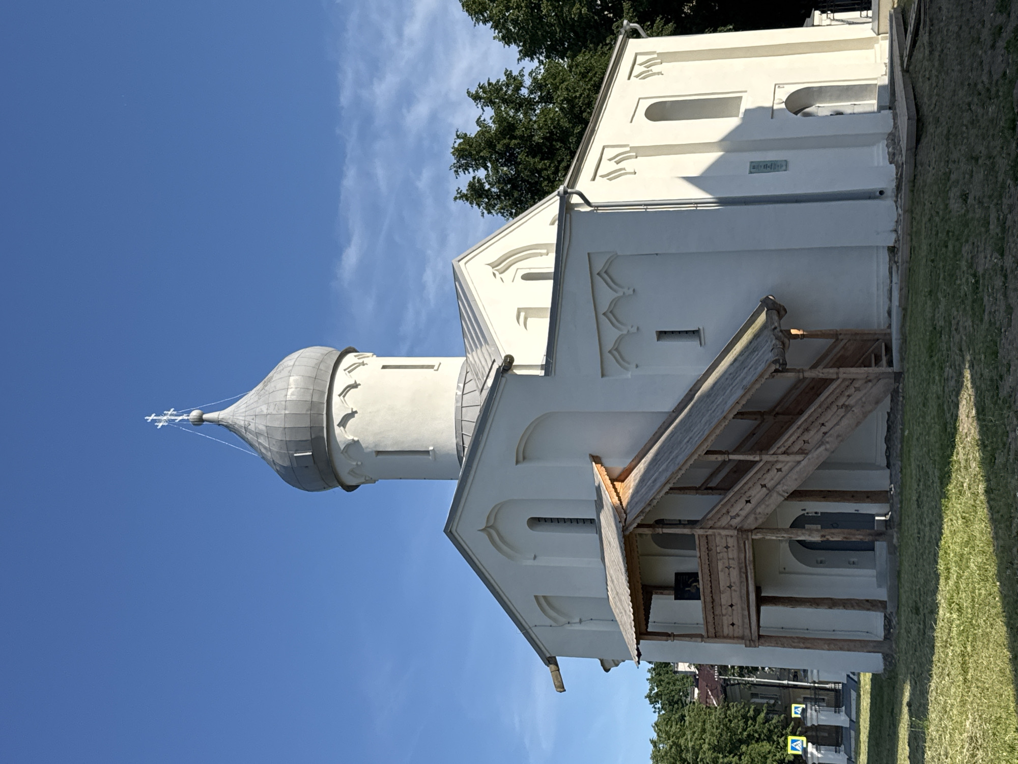 The image shows the Church of Saint George (Georgievskaya Tserkov) in Veliky Novgorod, Russia, on a clear, sunny day with a bright blue sky. The church is a white, stone building with a grey dome topped with a cross. A wooden staircase with a thatched roof leads up to an entrance on the left side of the building. The church is surrounded by a grassy lawn, with trees visible in the background. Two yellow traffic signs with pedestrian symbols are visible on the left side of the image, near a fence. The building appears to be of medieval architecture, with arched windows and decorative elements along the facade. The overall impression is one of historical preservation and serene beauty.