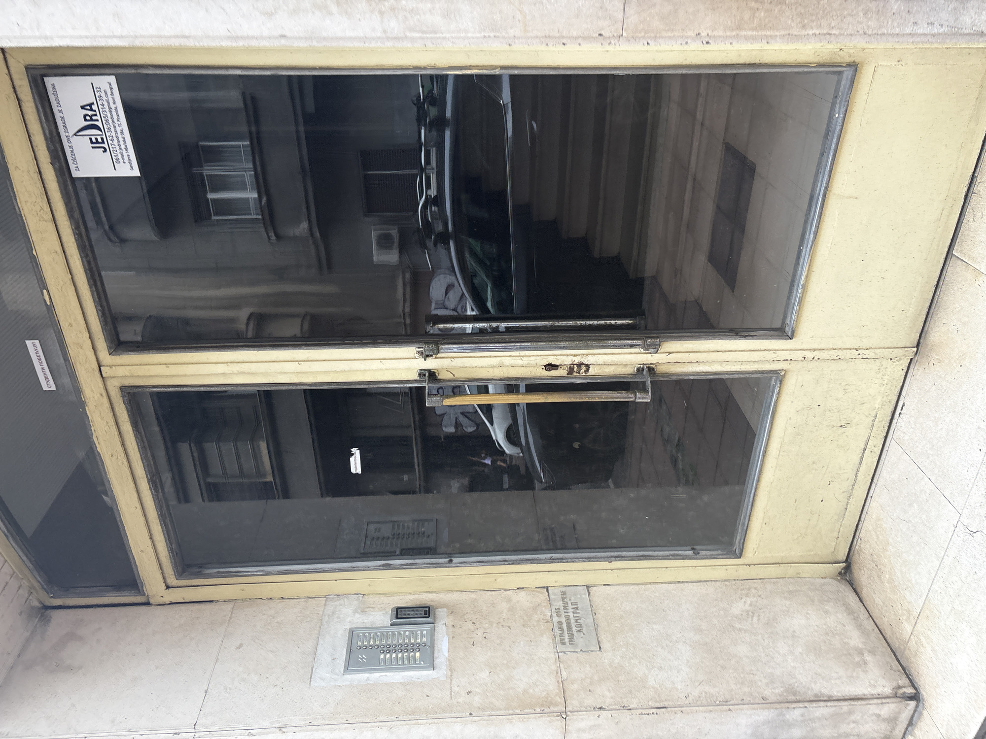 The image shows the entrance to a building in Belgrade, Serbia. The entrance has a double glass door with yellow-painted frames. On the left side of the entrance, there is a wall-mounted intercom system and a plaque with Serbian text. The door on the right has a sticker with the logo "JEDRA" and contact information, also in Serbian. Reflections in the glass doors show a dark-colored car, a set of stairs, and other architectural elements of the surrounding buildings. The overall scene appears to be an urban residential or commercial building entrance.