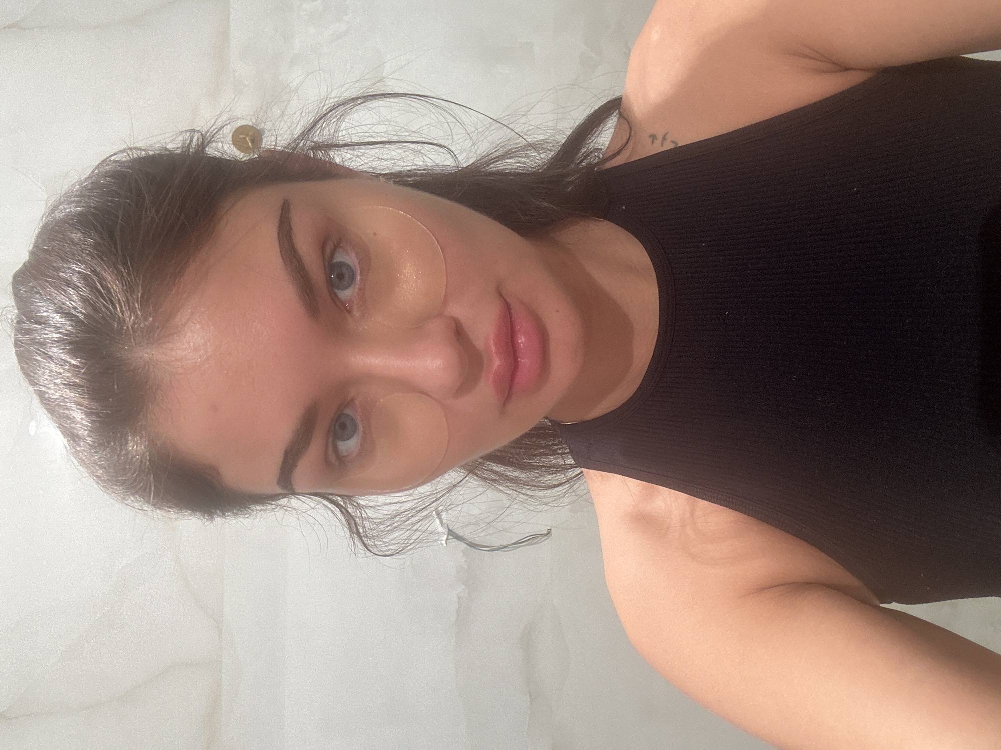 A woman is captured in a close-up shot. She has fair skin, dark hair, and blue eyes. She's wearing two translucent, circular patches on her face, possibly eye masks. She wears a black tank top. The background is a light-colored wall with a slightly textured surface. There is no indication of the location being Moscow, Russia.