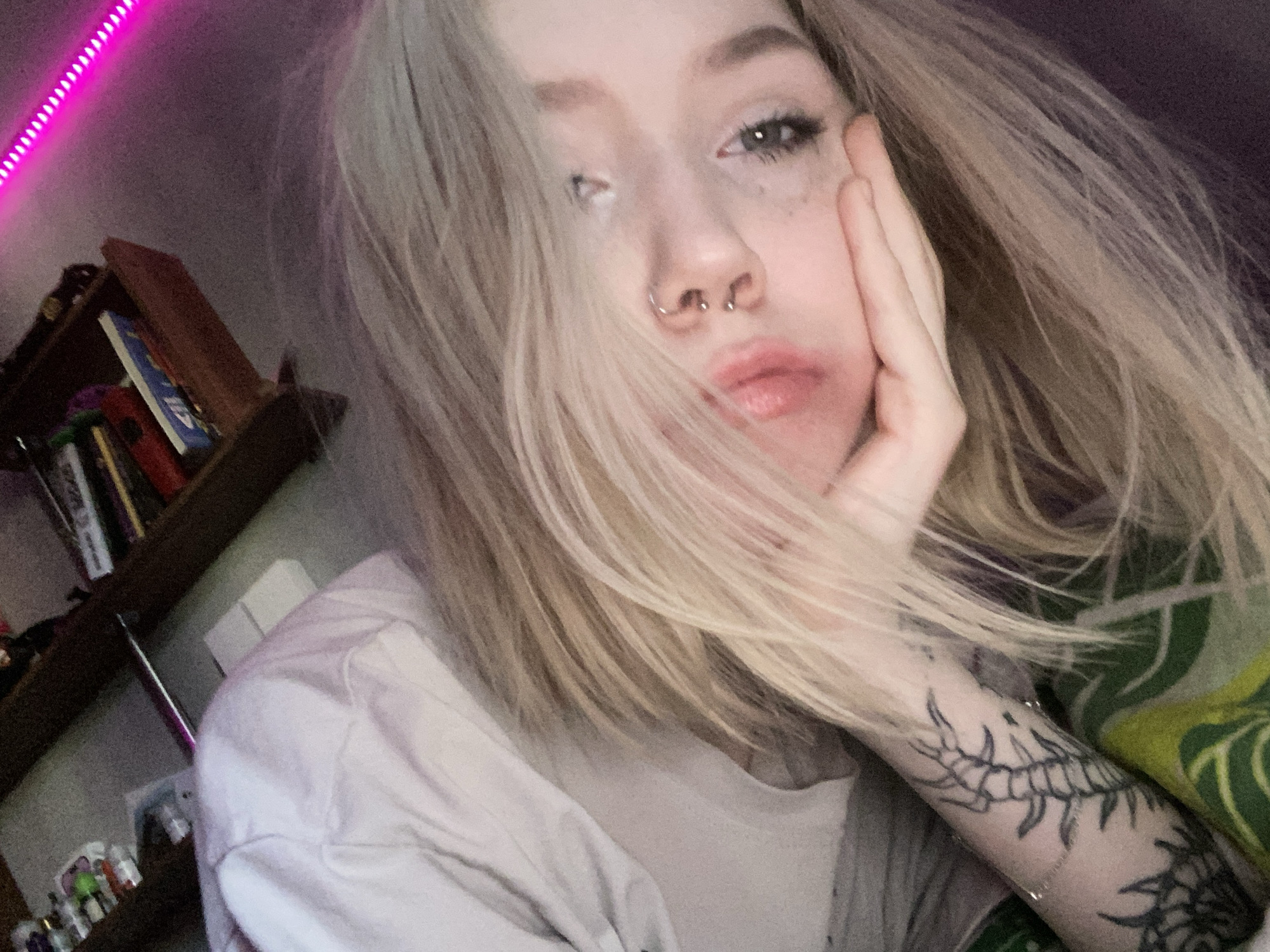 A close-up, tilted photograph depicts a fair-skinned individual, appearing female, with long blonde hair partially obscuring the left side of her face. She looks directly at the camera with blue or grey eyes, her right hand supporting her chin. Her nose features a silver septum ring and two silver studs as piercings. Dark mascara is visible on her lower eyelashes. On her left forearm, a prominent black tattoo resembling a segmented insect or centipede is visible, alongside another partial tattoo. A thin chain bracelet encircles her left wrist. She is wearing a light-colored top. The background indicates an indoor setting; a wooden shelf or cabinet containing assorted items, including small boxes or books, is visible in the upper right. A strip of bright pink LED lighting illuminates the upper right portion of the scene. Green and yellow patterned fabric is visible in the lower left, adjacent to what appears to be light-colored bedding or soft fabric. The specific location within Izhevsk, Russia, cannot be determined from the visual information provided.