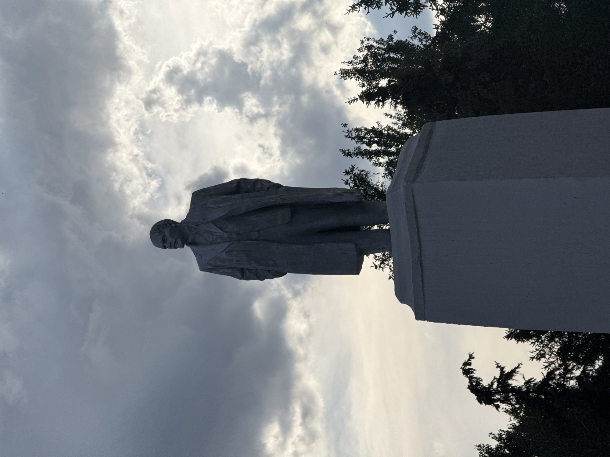 The media file depicts a statue of a man, likely a historical figure, standing on a pedestal. The statue is rendered in a grey, stone-like material and shows the man from the waist up, dressed in a suit and overcoat. He is facing left with his head turned slightly.
The scene is set outdoors against a dramatic sky filled with heavy, grey clouds, suggesting overcast or stormy weather. The lighting is dim, indicating it might be late afternoon or evening. To the right and upper left of the statue, dark green foliage from trees is visible, framing the sky and the monument.
The orientation of the image is such that the statue appears to be standing upright, with the pedestal at the bottom right of the frame and the statue extending upwards and to the left. The pedestal is a substantial, angular structure.
There is no visible text in the media file. The overall mood conveyed is somber and perhaps a bit imposing, due to the statue's presence and the heavy, cloud-filled sky. The location context provided, Masalovka, Russia, suggests a possible setting for such a monument. The image focuses on the statue and its immediate surroundings, highlighting the contrast between the man-made monument and the natural elements of the sky and trees.