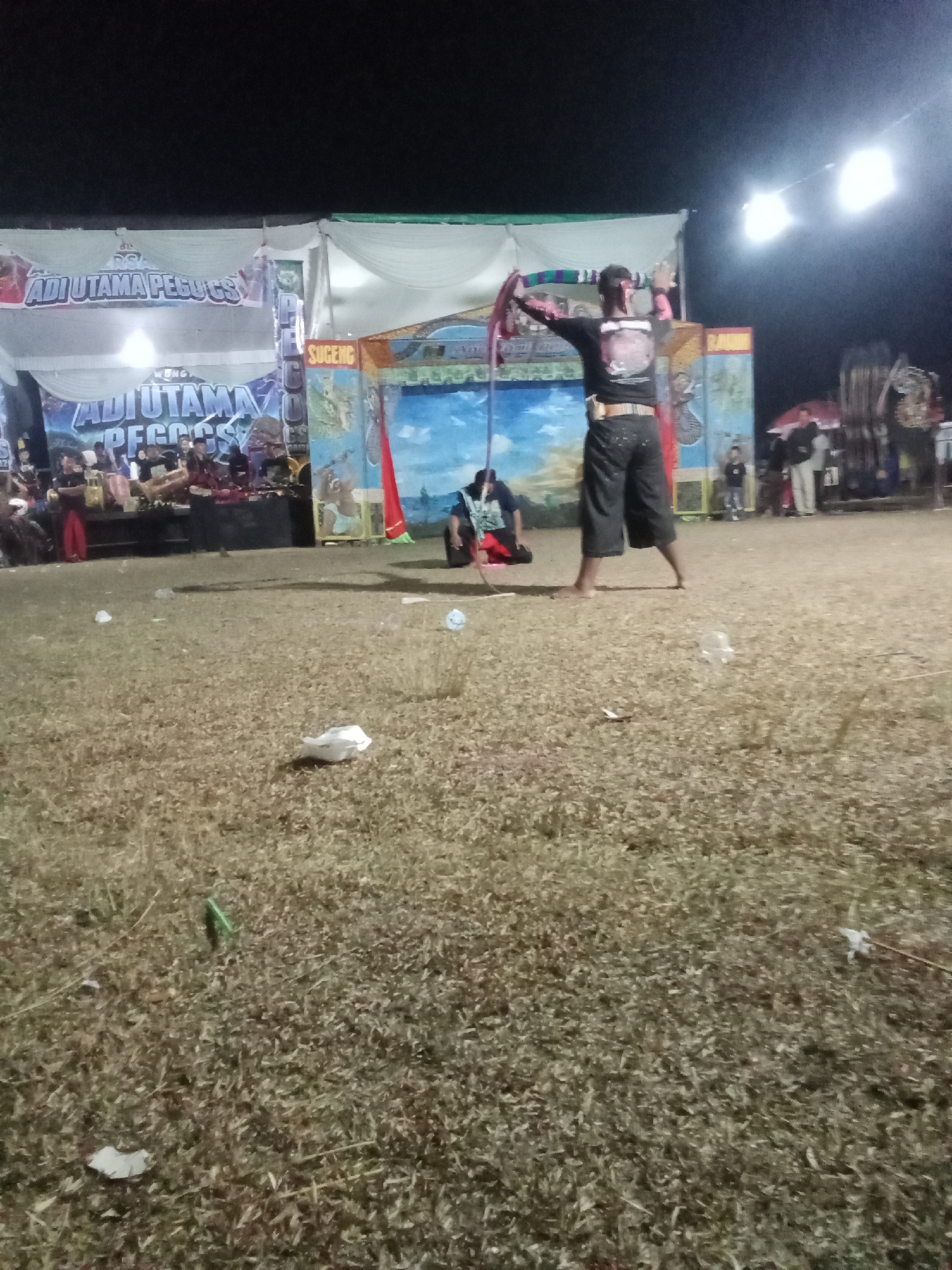 The image depicts an outdoor night scene in Sidodadi, Indonesia, likely at a cultural performance or festival. In the foreground, dry, sparse grass covers the ground, with scattered pieces of litter. In the mid-ground, a male performer, dressed in dark pants and a black t-shirt with a graphic design, stands with his back to the camera. He is holding a colorful, striped cloth or scarf in his outstretched hands, with his arms raised. To his left, another person is crouched or sitting on the ground. Behind the performer, a stage is set up with banners and a backdrop featuring a painted sky and landscape. The banner visible at the top left reads "ADI UTAMA PEGUS." Further back, there are other figures, some appearing to be spectators and performers, as well as equipment like speakers and musical instruments. Bright lights illuminate the area from above, suggesting it is an organized event.