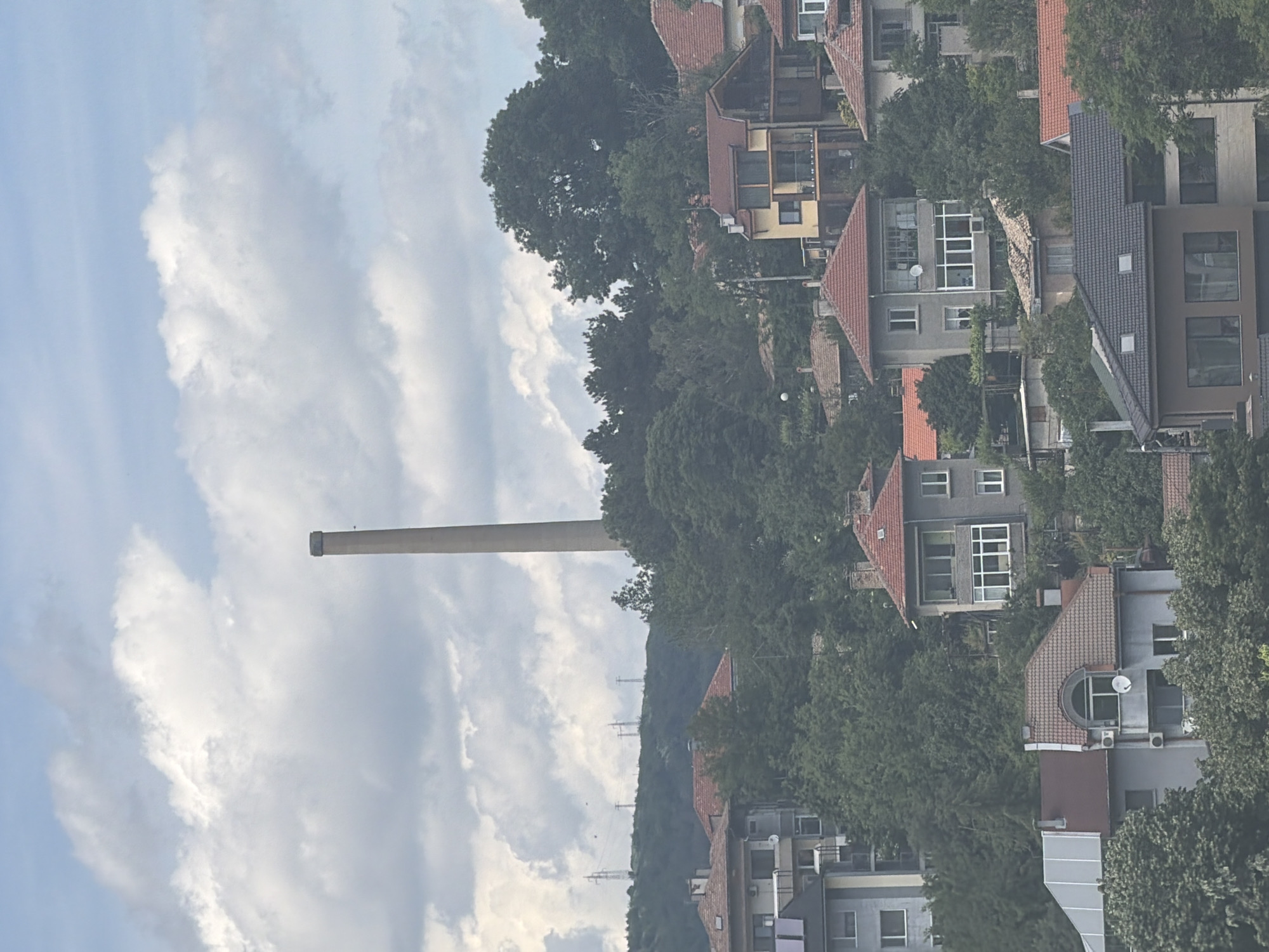 This image captures a panoramic daytime view of a residential area in Shumen, Bulgaria, set against a backdrop of hills and a cloudy sky. The most prominent object is a very tall, slender, light-colored cylindrical chimney, likely industrial, which stands vertically in the left-center of the frame against the overcast sky.
The scene features numerous multi-story residential buildings with varying architectural styles. Most houses have distinct red or grey tiled roofs, with facades in shades of grey and beige. Many buildings include balconies, white-framed windows, and some are equipped with satellite dishes. Dense green trees and other vegetation are abundant, covering the hillsides and interspersed among the houses, suggesting a lush, natural environment integrated with the urban development.
In the distant background, a series of electricity transmission towers can be observed ascending a tree-covered hill. The sky is largely covered with large, diffuse white and grey clouds, indicating overcast weather during daylight hours, likely morning or afternoon due to the soft, even lighting. No people or specific activities are discernible in the static landscape. There is no visible text present in the image.