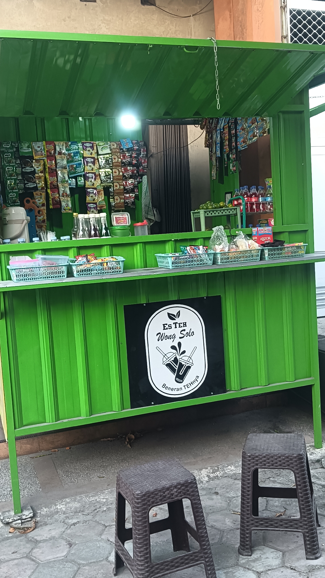 The image depicts an outdoor stall in Sragen, Indonesia, likely a small food or beverage vendor. The stall is painted bright green and features a corrugated metal roof. Various packaged goods, such as snacks and drinks, are displayed on shelves. A prominent sign with a black background and white text reads "Es Teh Wong Solo" and features an illustration of two iced tea glasses. In front of the stall, two brown plastic stools are placed on a cobblestone-like ground. The overall scene suggests a casual commercial establishment operating in an urban or semi-urban environment.