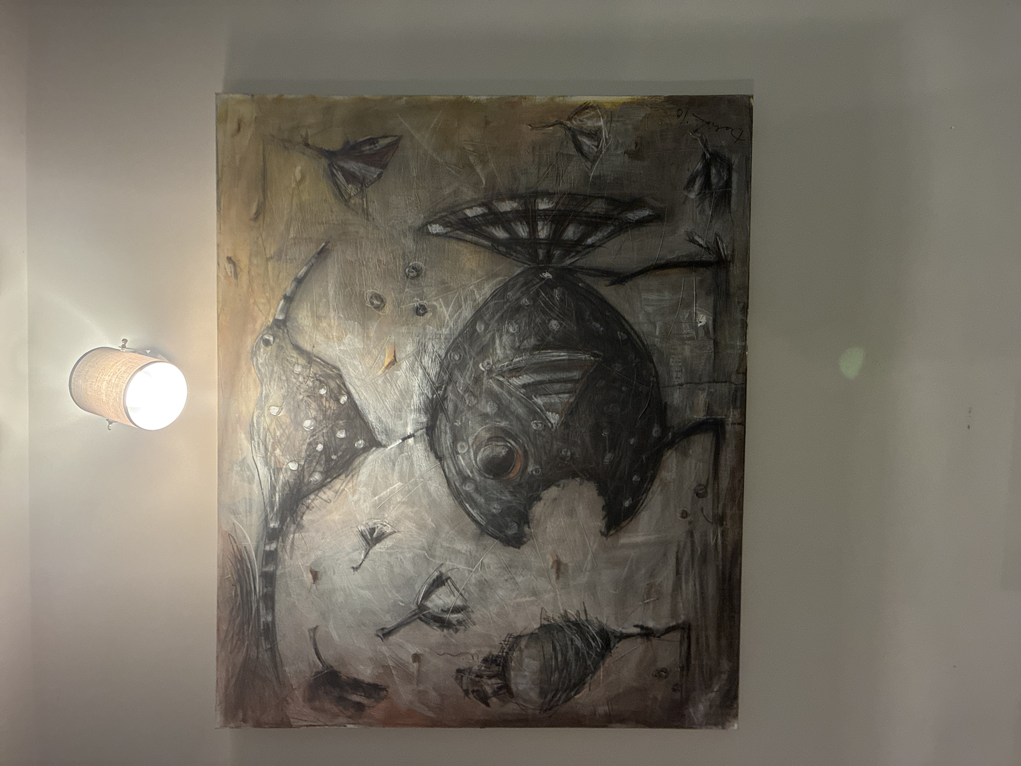 An abstract and surreal painting dominates the center of the image, hanging on a plain, light-colored wall. A wall-mounted lamp with a fabric shade illuminates the artwork from the left side.
The painting features dark, organic forms against a lighter, textured background of grays, browns, and muted earth tones, with visible brushstrokes and scratches. A prominent, large, dark, and rounded shape, reminiscent of a stylized fish or a fantastical deep-sea creature, occupies the central area. This central figure has a circular, eye-like detail and a distinct, multi-layered structure resembling a fin or a crown on its top. Surrounding this main form are several smaller, dark, and elongated shapes that evoke similar creature-like or insectoid figures, floating within the textured background.
In the upper right corner of the painting, faint, handwritten text or a signature is visible, appearing to read "Damon '10" or similar, indicating the artist and the year 2010. The setting is indoors, with the artificial light suggesting an evening or nighttime display of the artwork. The overall impression of the painting is mysterious and otherworldly.