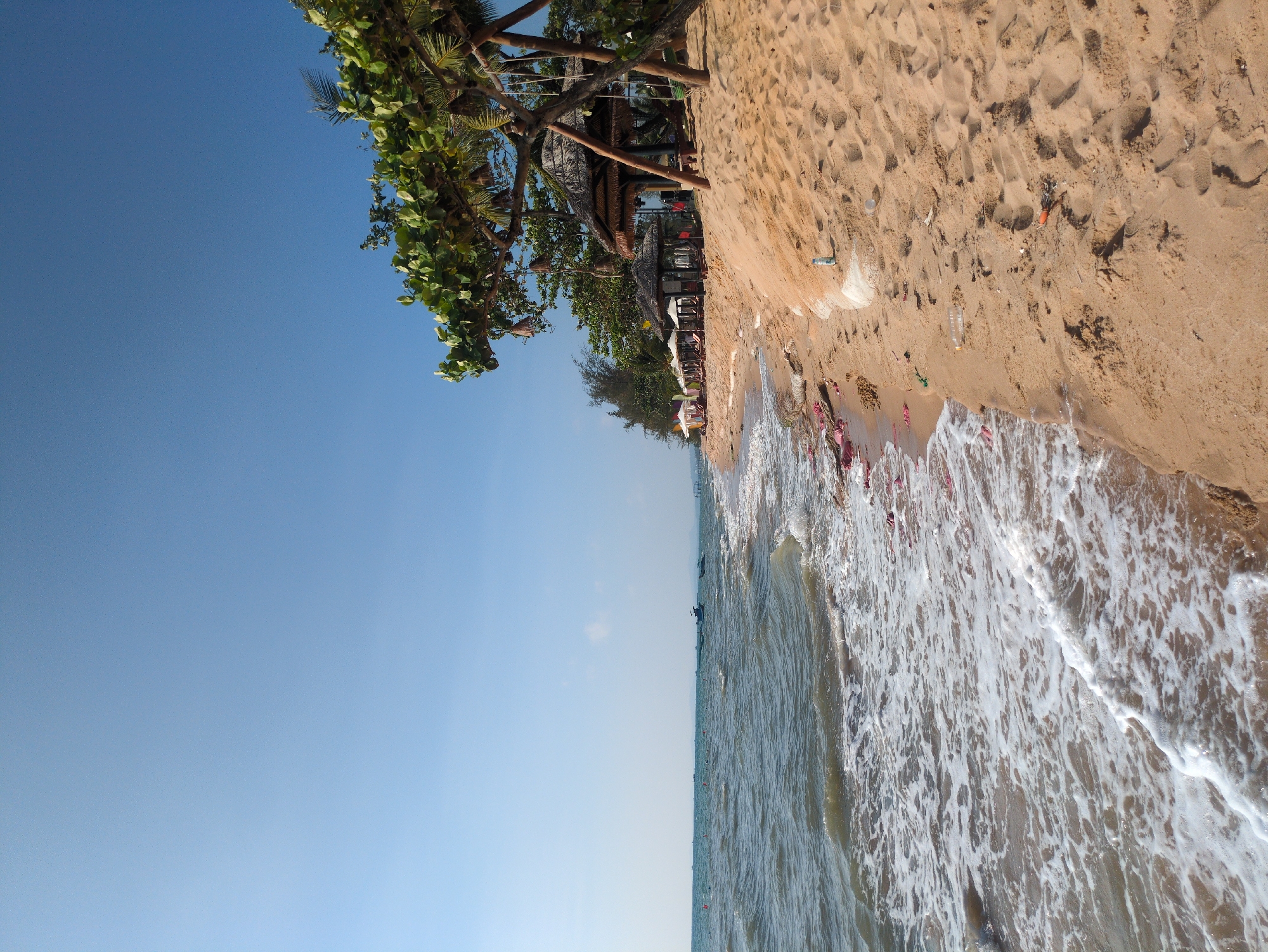 The image shows a beach scene in Phú Quốc, Vietnam. The view is oriented vertically, with the bright blue sky dominating the upper left portion. A tree with green leaves is visible in the upper left corner, near some beach huts. The right side of the image is occupied by the sandy beach, littered with debris. The ocean borders the sand, with white waves crashing on the shore.