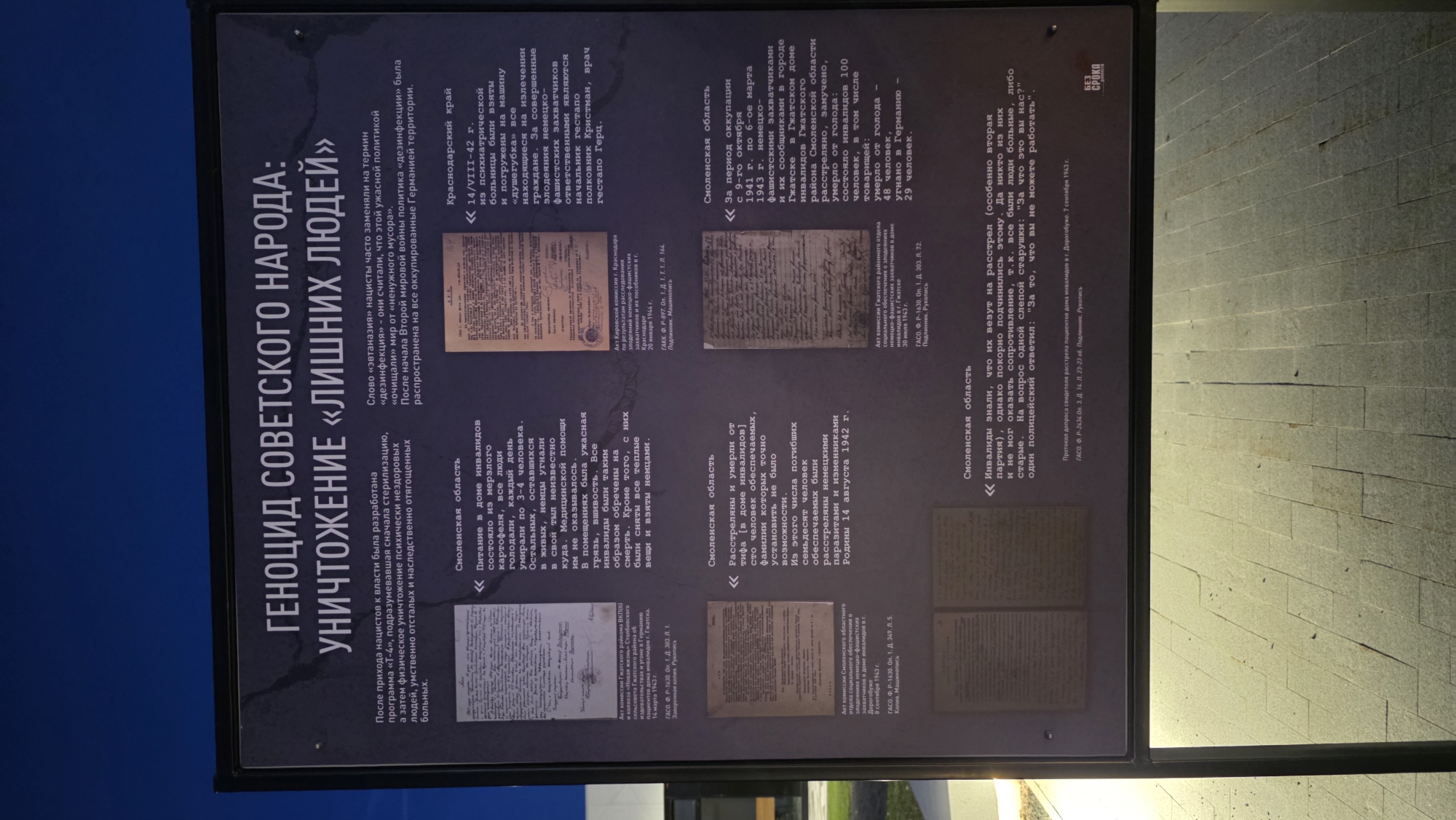 The image shows an outdoor information panel displaying information about the genocide of the Soviet people during World War II, specifically focusing on the extermination of "superfluous people" (those deemed undesirable by the Nazi regime). The location is likely a memorial or historical site. The panel contains text in Russian and reproductions of historical documents related to the persecution and extermination of disabled people, mentally ill individuals, and others deemed unfit by the Nazi regime in occupied territories of the Soviet Union, such as the Smolensk and Krasnodar regions. The documents include reports, testimonies, and official communications detailing atrocities committed by German forces. The panel is placed in an outdoor location with a paved area in the background.