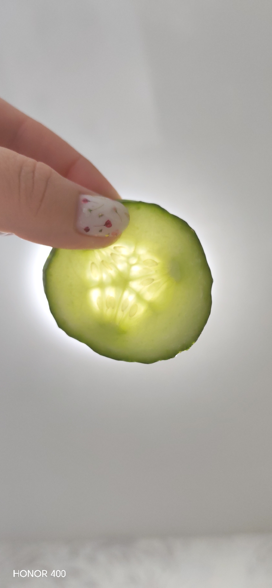 A hand with a decorated fingernail is holding up a slice of cucumber, backlit. The slice of cucumber is round with seeds visible in the center. The background is a light, blurry white. The lower left corner of the image has the text "HONOR 400". There is no evidence this image was taken in Noviy, Russia.