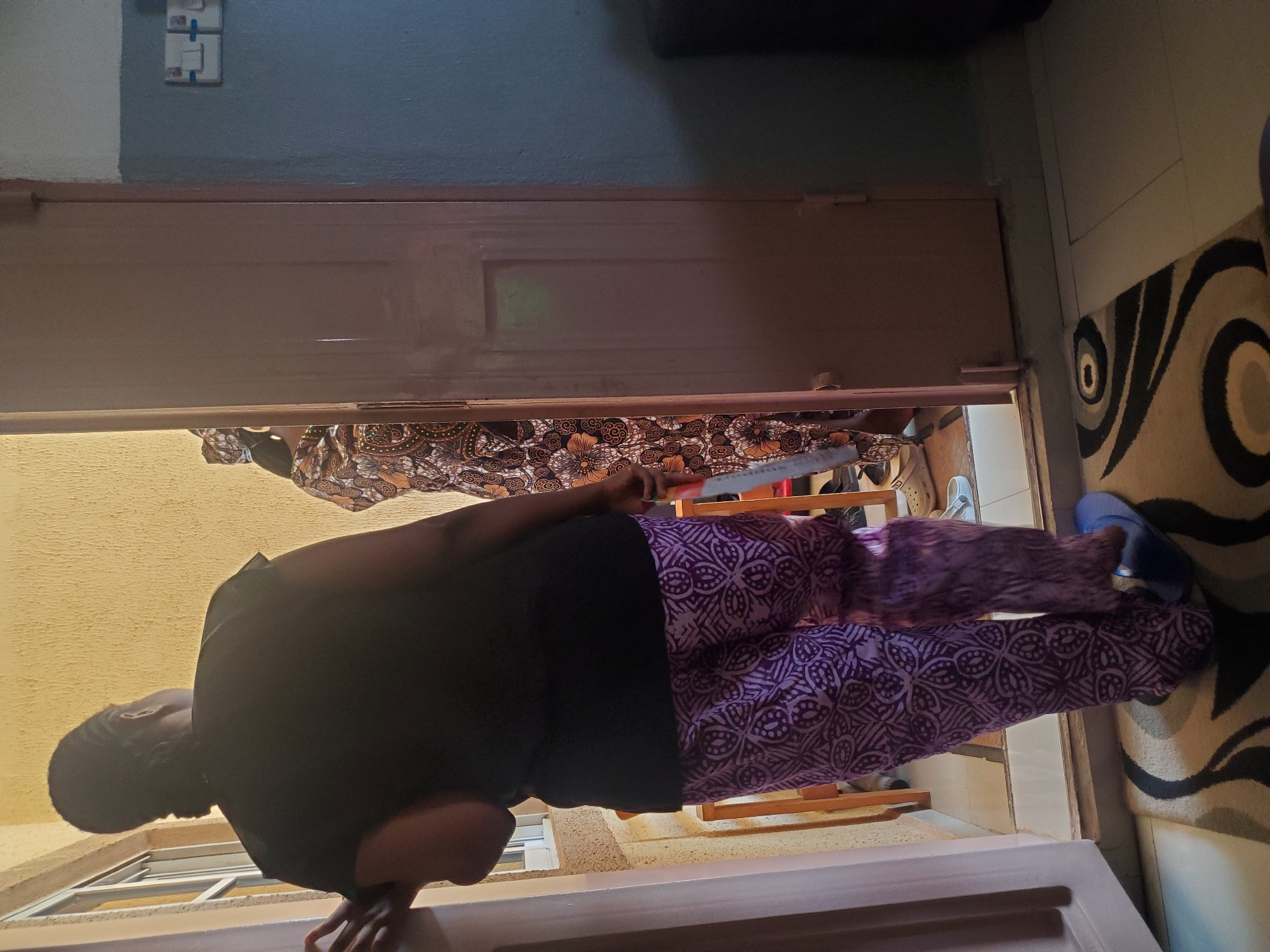This image captures an interior scene in Sabon Tasha, Nigeria, looking through an open doorway into an adjacent space. In the immediate foreground, a person is visible from the back, wearing a black short-sleeved top and purple pants with intricate white/light purple geometric patterns. This individual has dark, braided hair pulled back and is wearing blue slip-on sandals. Their left hand rests on the light-colored doorframe.
Through the open doorway, a second person is partially visible, also from the back. This individual wears a long, patterned garment in shades of brown, orange, and black, complemented by a matching head covering. Their right hand holds a slender, light-colored object, possibly a broom or brush handle, with a red accent near the top.
The interior floor features a beige carpet with a prominent black and grey abstract swirling pattern, transitioning to light-colored tiles further back. To the right of the doorway, a two-tone wall is visible, with its lower section painted a dark teal-grey and the upper section a lighter hue. Two white, rectangular electrical outlets or switches are mounted on the upper part of this wall. A dark, possibly black, object is partially visible on the far right, suggesting furniture.
Beyond the doorway, the adjacent space reveals a textured, light yellow wall and a window with white frames on the left. A low wooden railing or barrier is present, and several pairs of footwear, including white clogs and sandals, are visible on the tiled floor. The overall lighting suggests ambient daylight entering from the exterior.