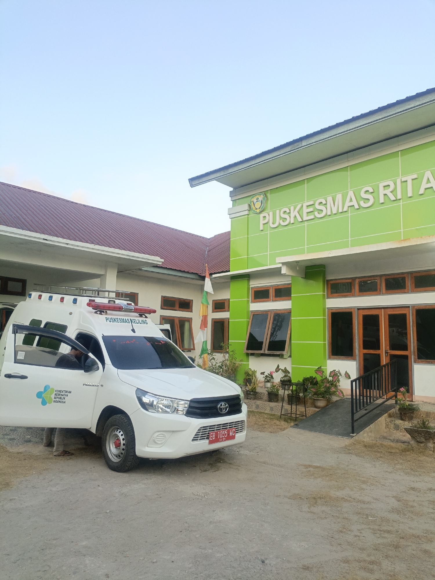 An exterior, eye-level, medium shot captures a white ambulance parked in front of a modern building with a lime green facade. The ambulance is a Toyota Hilux, and its front left door is open, revealing a person seated in the driver's seat. The side of the ambulance displays the logo for "KEMENTERIAN KESEHATAN REPUBLIK INDONESIA" (Ministry of Health of the Republic of Indonesia) and the words "PUSKESMAS KELILING" (Mobile Health Center). Above the ambulance, the building's green facade is emblazoned with the words "PUSKESMAS RITA" (Rita Health Center). To the left of the ambulance, a building with a red roof is partially visible. The foreground consists of a dirt and gravel surface. In the background, a clear blue sky is visible. The scene is set in Solor Barat, Indonesia.