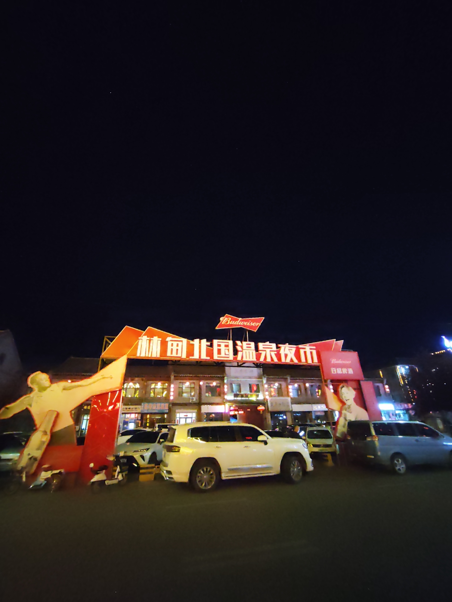 The image shows a nighttime street scene in Lindian, China, with a focus on a lively night market. A large, illuminated sign with red lettering in Chinese characters, reading "林甸温泉夜市" (Lindian Hot Spring Night Market), spans across the top of a building. The Budweiser logo is prominently displayed on the signage. To the left, a large, yellow, stylized figure, possibly a statue or cutout, is illuminated against the dark sky. In front of the building, several vehicles are parked, including a white SUV, a white scooter, and a blue minivan. A billboard featuring a portrait of a young woman is visible on the right side of the building. The street is paved, and the surrounding buildings are lit with various lights, indicating commercial activity. The sky above is dark and clear, with a few visible stars.
