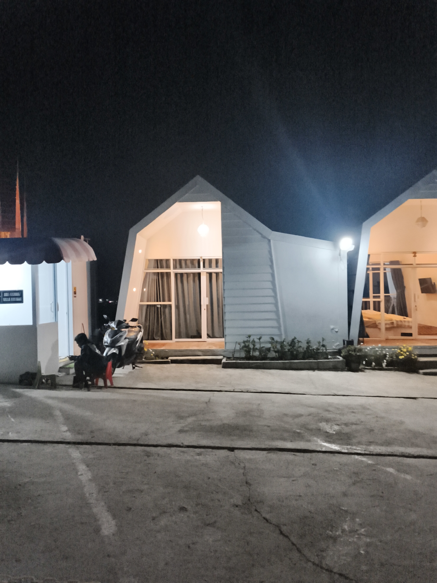 At night in Dieng Kulon, Indonesia, a residential building with a distinctive angular white facade is illuminated. Two modern, minimalist structures are visible. The left structure is a smaller, white kiosk-like building with a striped awning and a sign. In front of it, a person is seated on a red stool next to a dark motorcycle. The larger structure to the right is a two-story building with large glass windows and doors, revealing a bedroom interior with a bed and a television. The exterior walls of this building are white and feature horizontal paneling on one side. Both buildings are set on a concrete surface, with a small garden bed containing plants and yellow flowers in front of the right structure. The sky is dark, indicating nighttime.