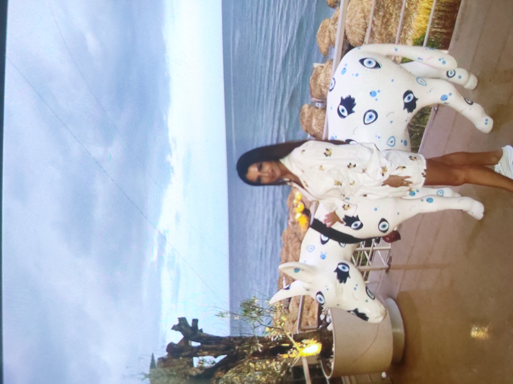 The image shows a woman in a light-colored dress posing with a decorative animal sculpture resembling a donkey or horse. The sculpture is white with various blue and black symbols, including evil eye motifs. The woman has dark hair and is smiling towards the camera.
The background includes a view of the sea and sky. A tree or shrub in a pot is also visible. Based on visual cues, the location appears to be near the Black Sea coast. The presence of decorative animal sculptures and the coastal environment suggest this might be in Tsarevo, Bulgaria. Hay bales are visible in the background as well. The picture seems to be taken outdoors on a sunny day.